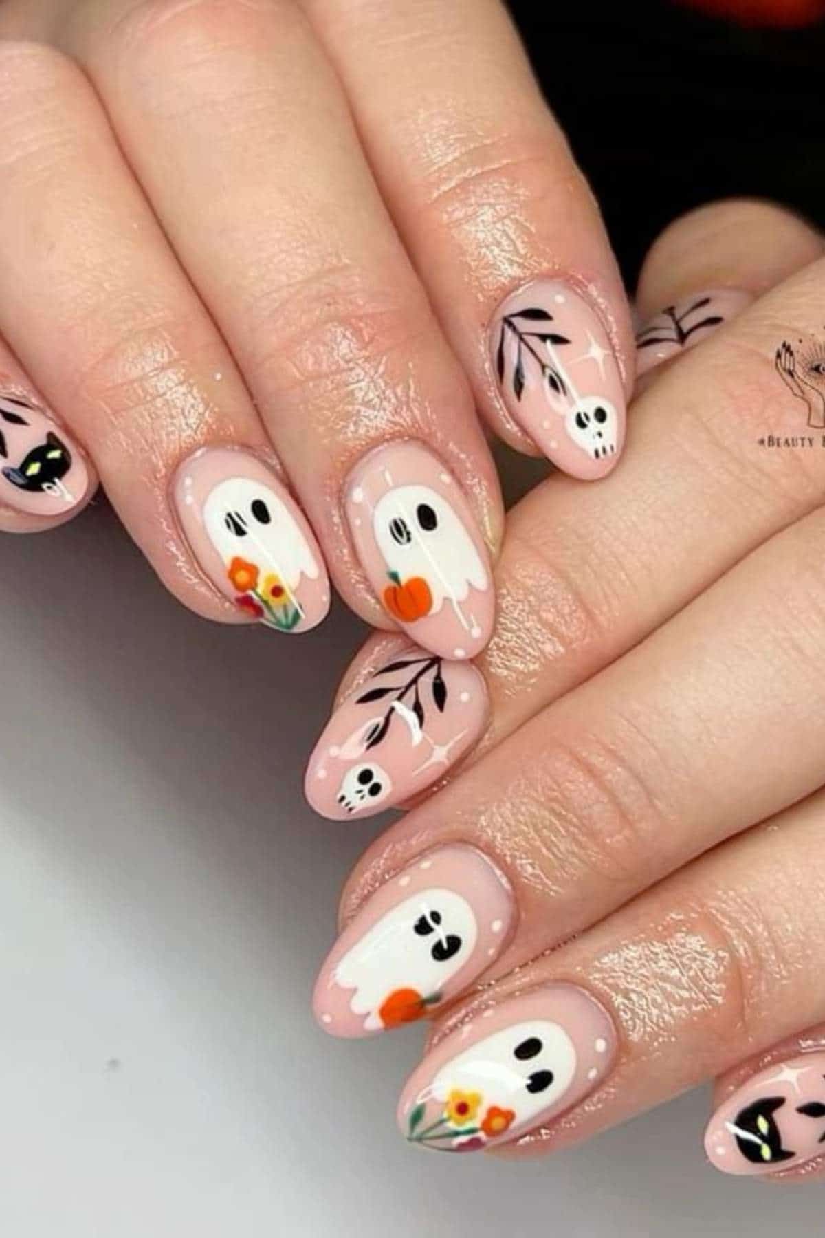 Hands with almond-shaped nails featuring Halloween-themed nail art, including ghosts, skulls, black leaves, and small colorful flowers on a nude pink base.