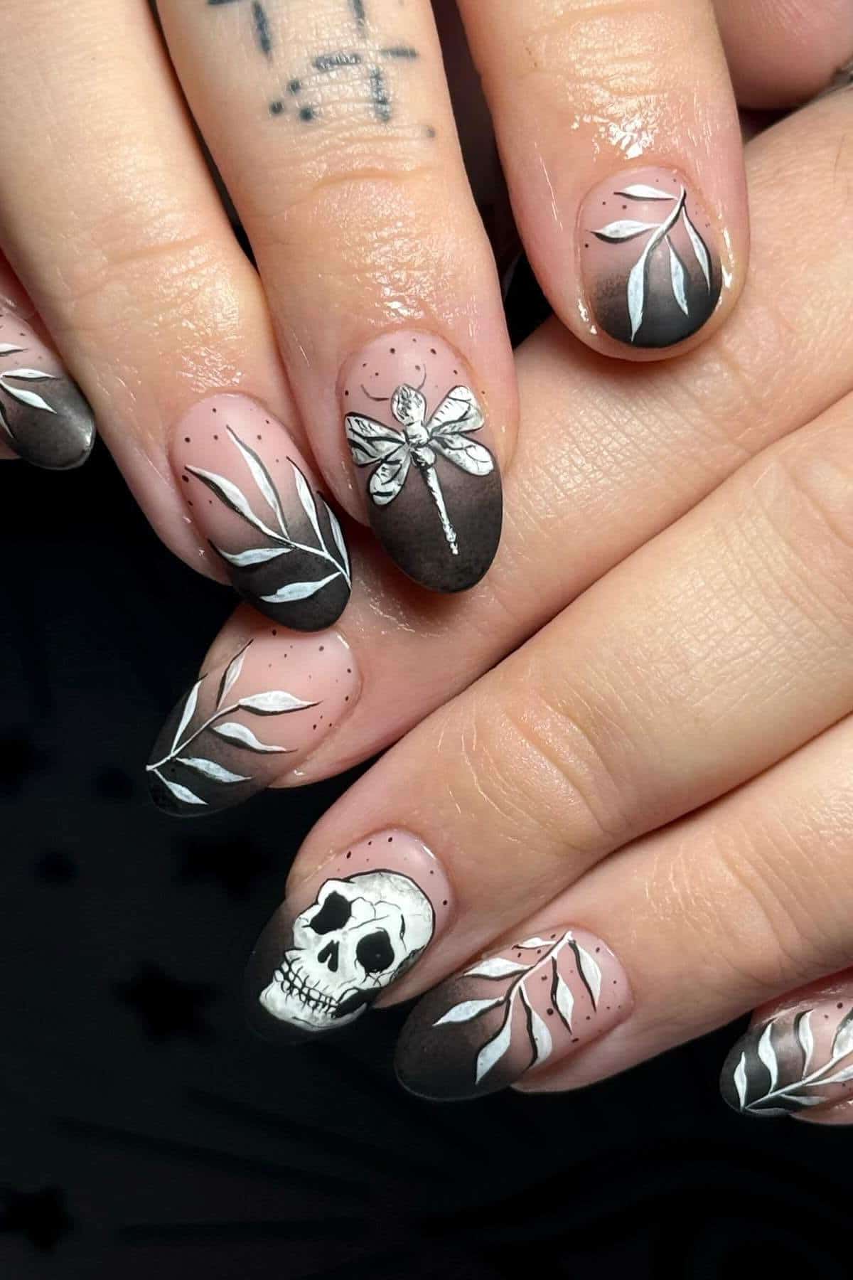 Close-up of hands with almond-shaped nails featuring black and nude gradient, white skull, dragonfly, and leaf designs. Some fingers have small dot accents and visible hand tattoos.