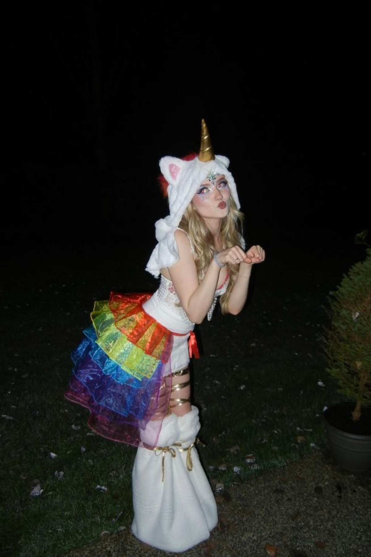 Person dressed in a unicorn costume with a rainbow skirt, unicorn horn, and white boots, posing outdoors at night with hands held up in a playful manner.