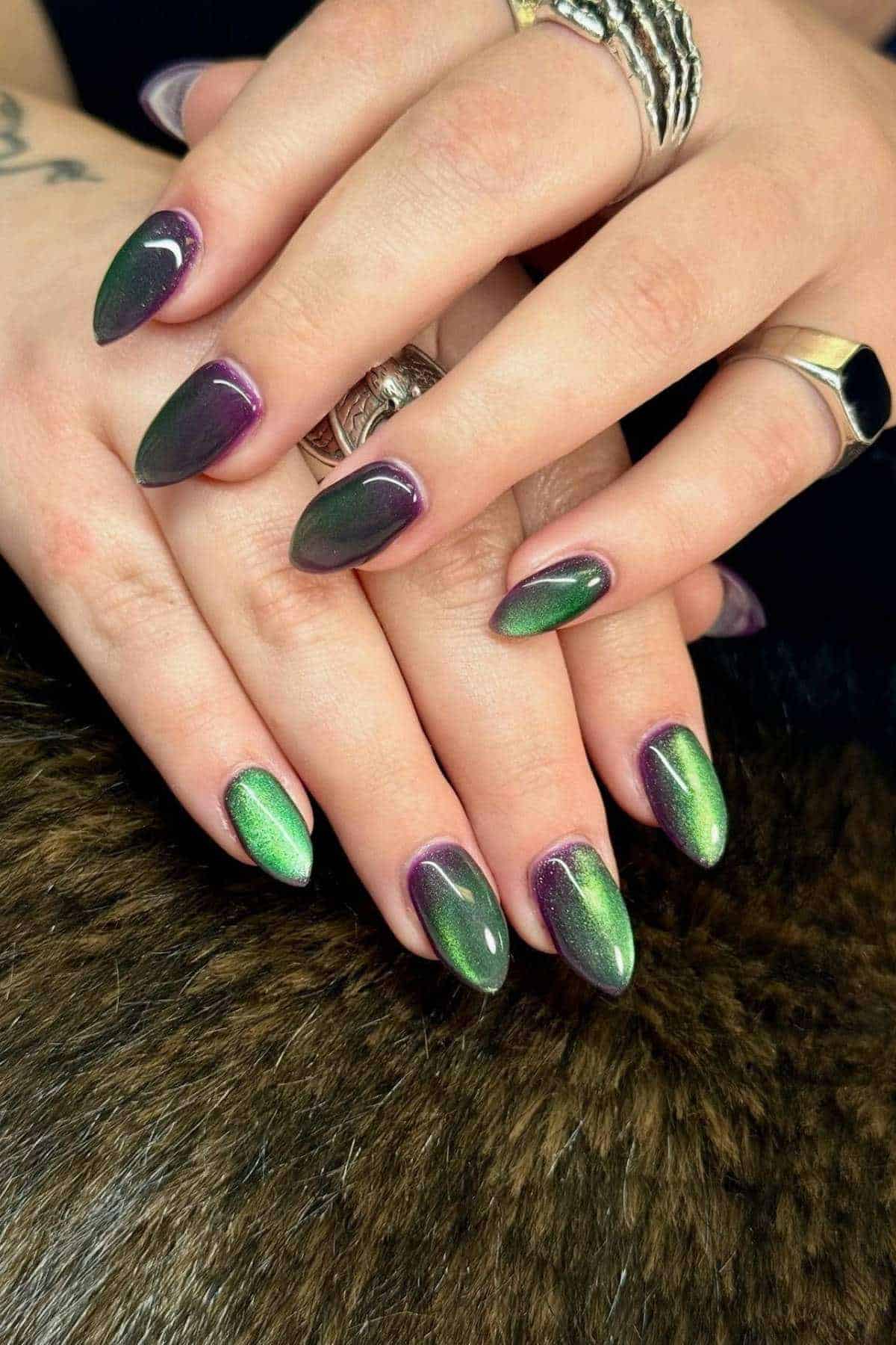 Hands with almond-shaped nails featuring a green and purple cat-eye effect manicure, resting on a dark, textured surface. Multiple silver rings are worn on the fingers.
