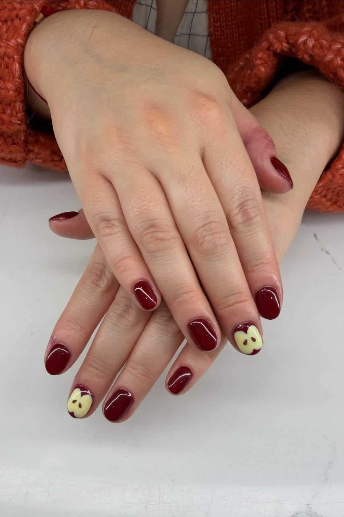 Two hands with short, rounded nails painted dark red, with the ring fingernails featuring a cream-colored base and dark red heart-shaped designs near the tips.