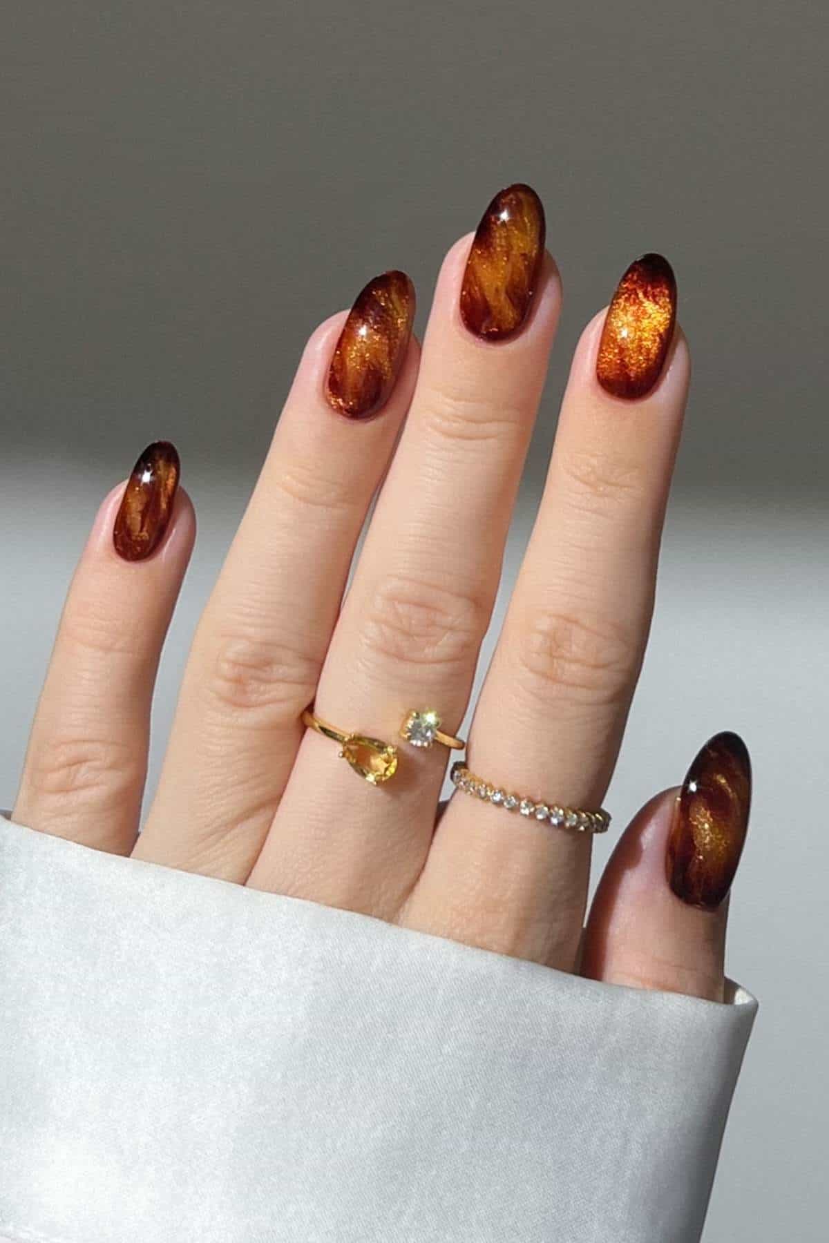 A hand with almond-shaped nails painted in a glossy, amber-brown gradient design, wearing two gold rings, shown against a neutral background.