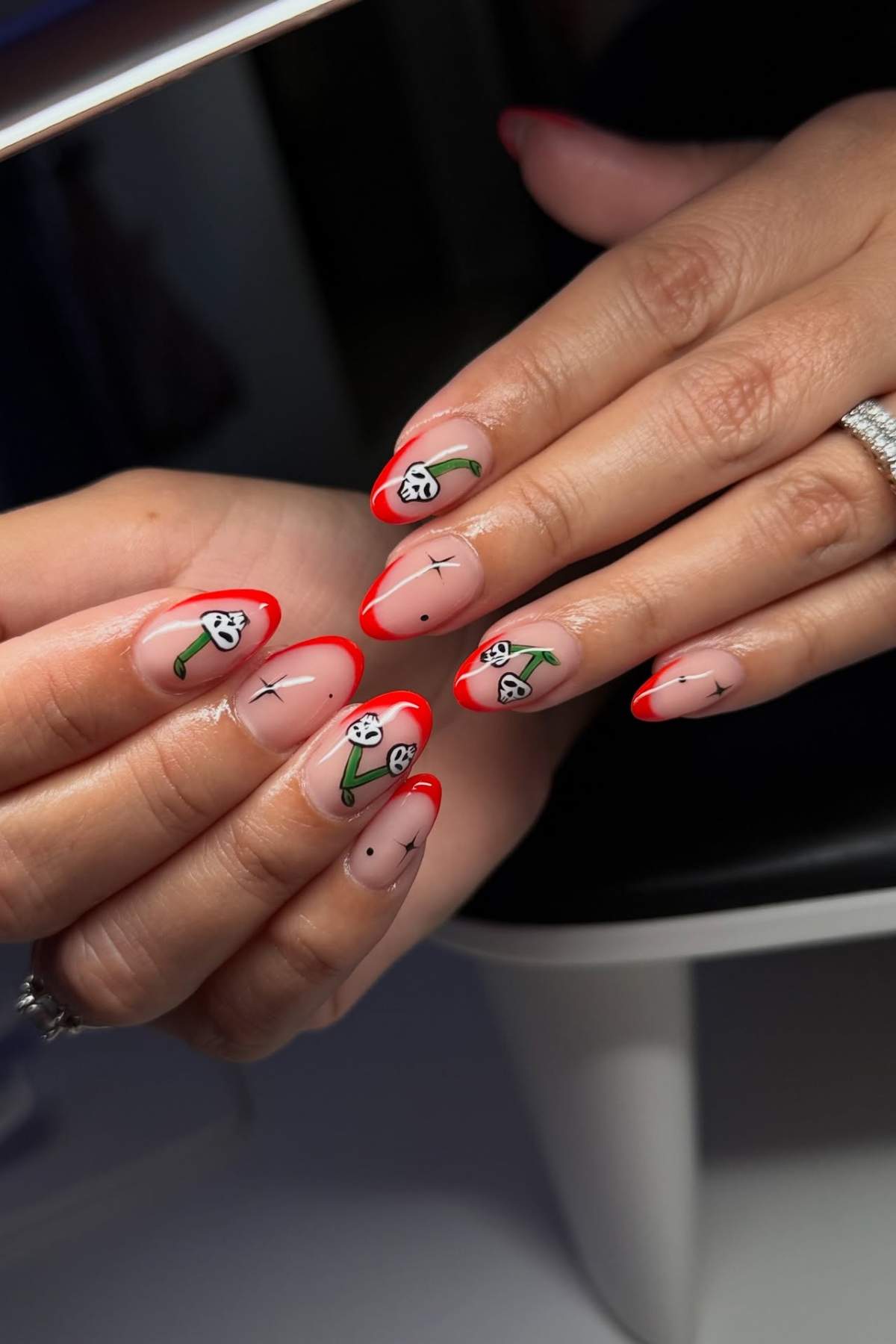 Close-up of hands with almond-shaped nails featuring red French tips and playful cherry and smiley face nail art on a nude base.