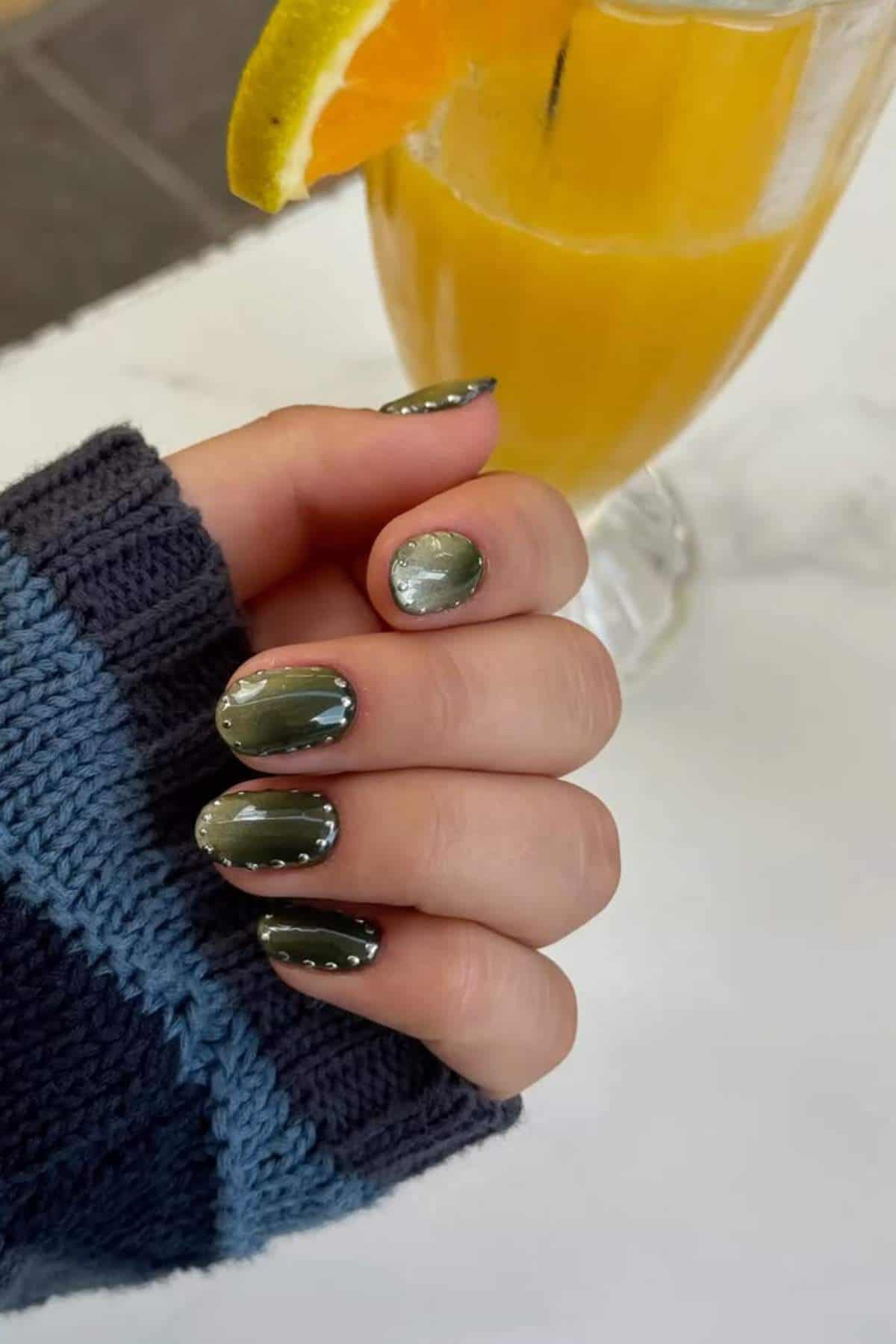 A hand with green and silver nail art holds a glass of orange beverage garnished with a slice of orange; the person is wearing a blue knit sweater.