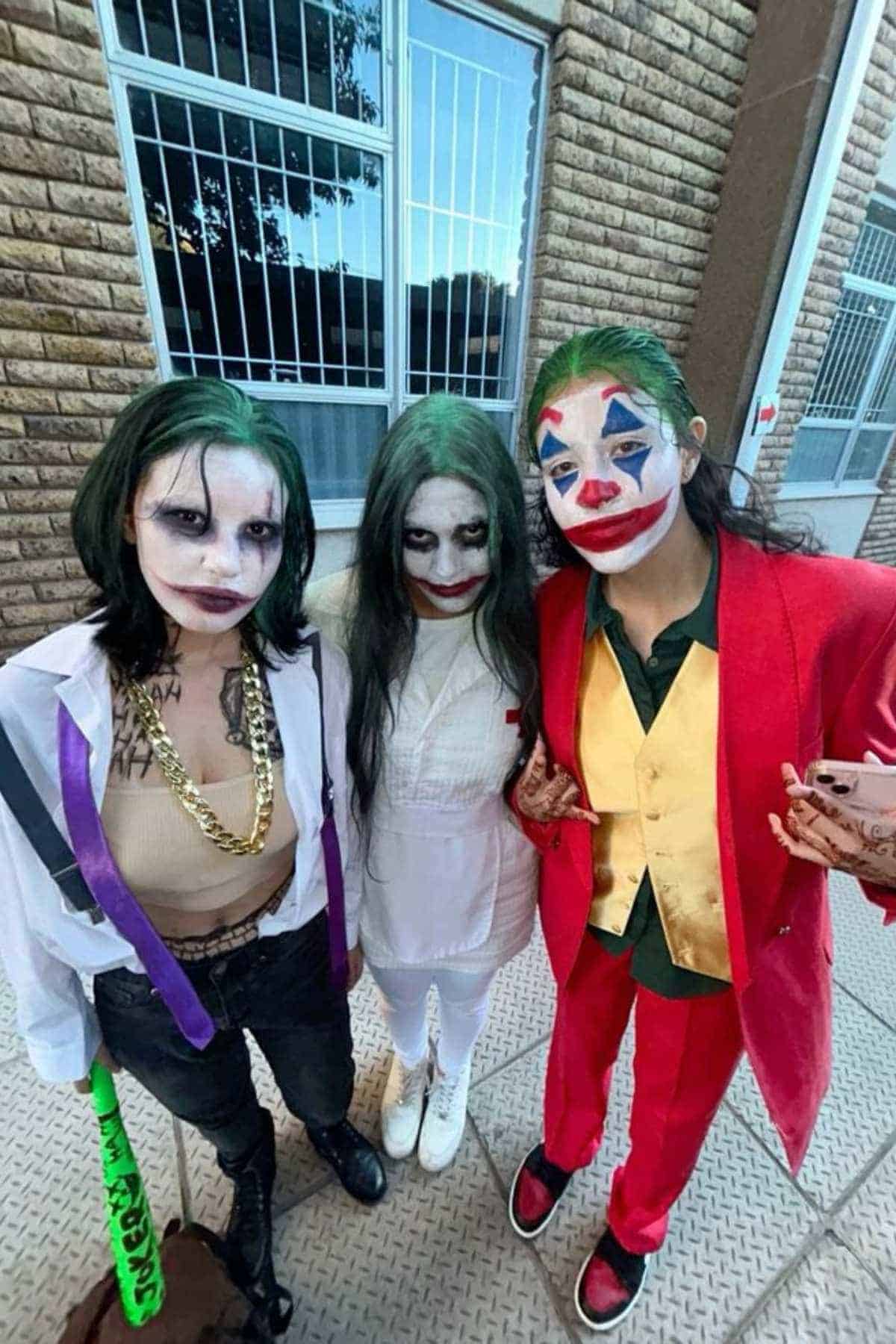 Three people dressed as different versions of the Joker character stand together outside near a brick wall and barred windows.