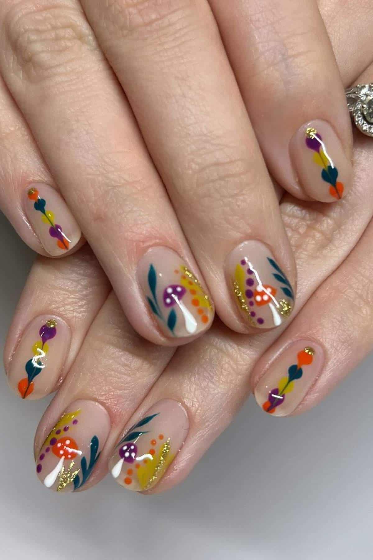 Fingernails with a clear base are decorated with colorful abstract designs, including flowers, leaves, mushrooms, and gold glitter accents.