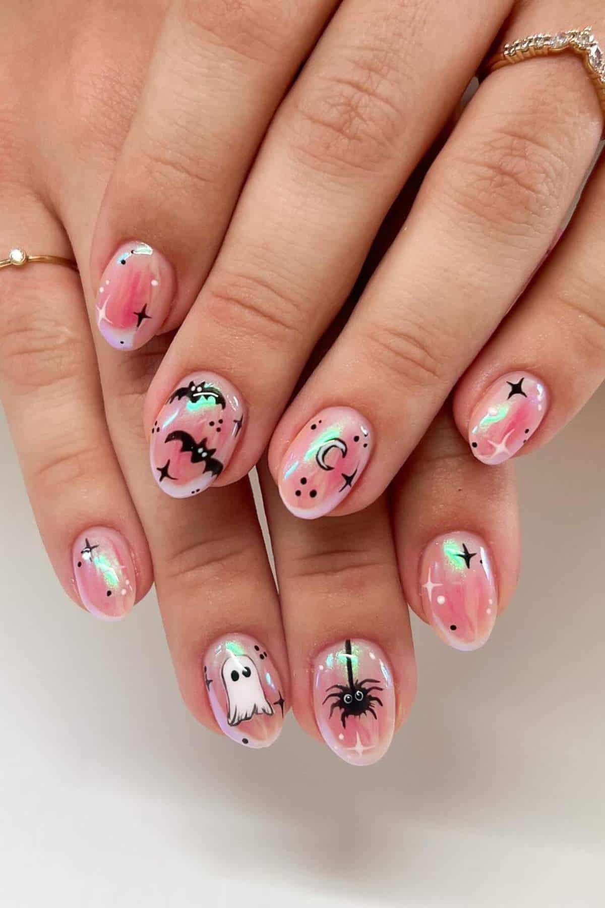 Hands with short, rounded nails painted with a pink base and decorated with Halloween-themed designs like ghosts, bats, spiders, stars, and moons.