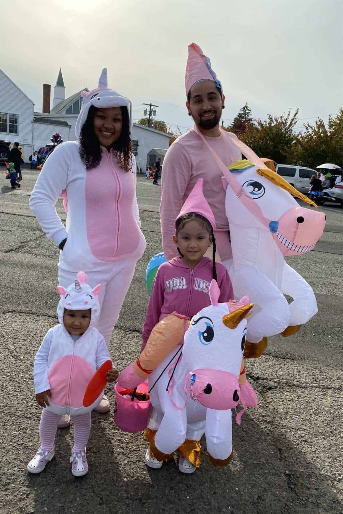 A family of four in unicorn-themed costumes, standing outdoors on pavement. Two adults and two children are smiling at the camera, with houses and trees in the background.