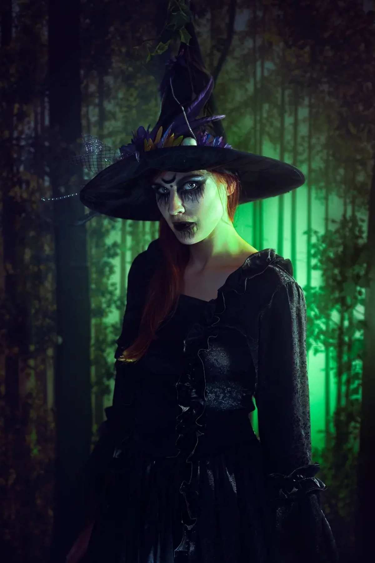 Person in black witch costume with face paint and pointed hat stands in a dark forest with green light in the background.