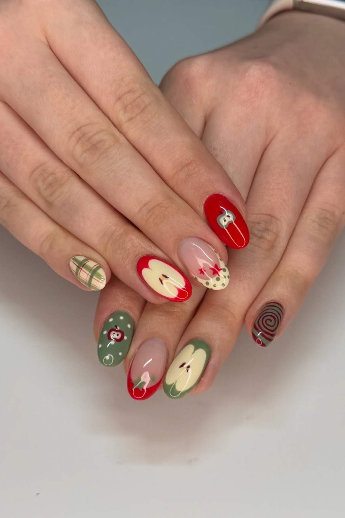Hands with long oval nails featuring apple, plaid, swirl, and abstract designs in red, green, and beige tones.