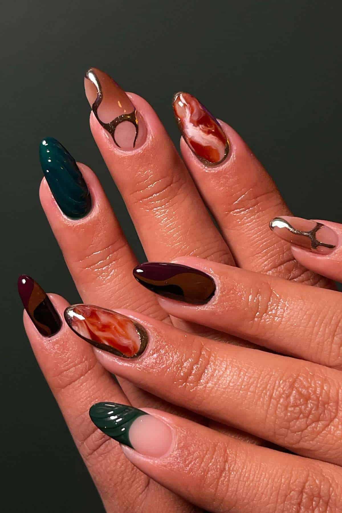 Hands with long almond-shaped nails, featuring a mix of dark green, burgundy, nude, and marbled designs with metallic outlines on a dark background.
