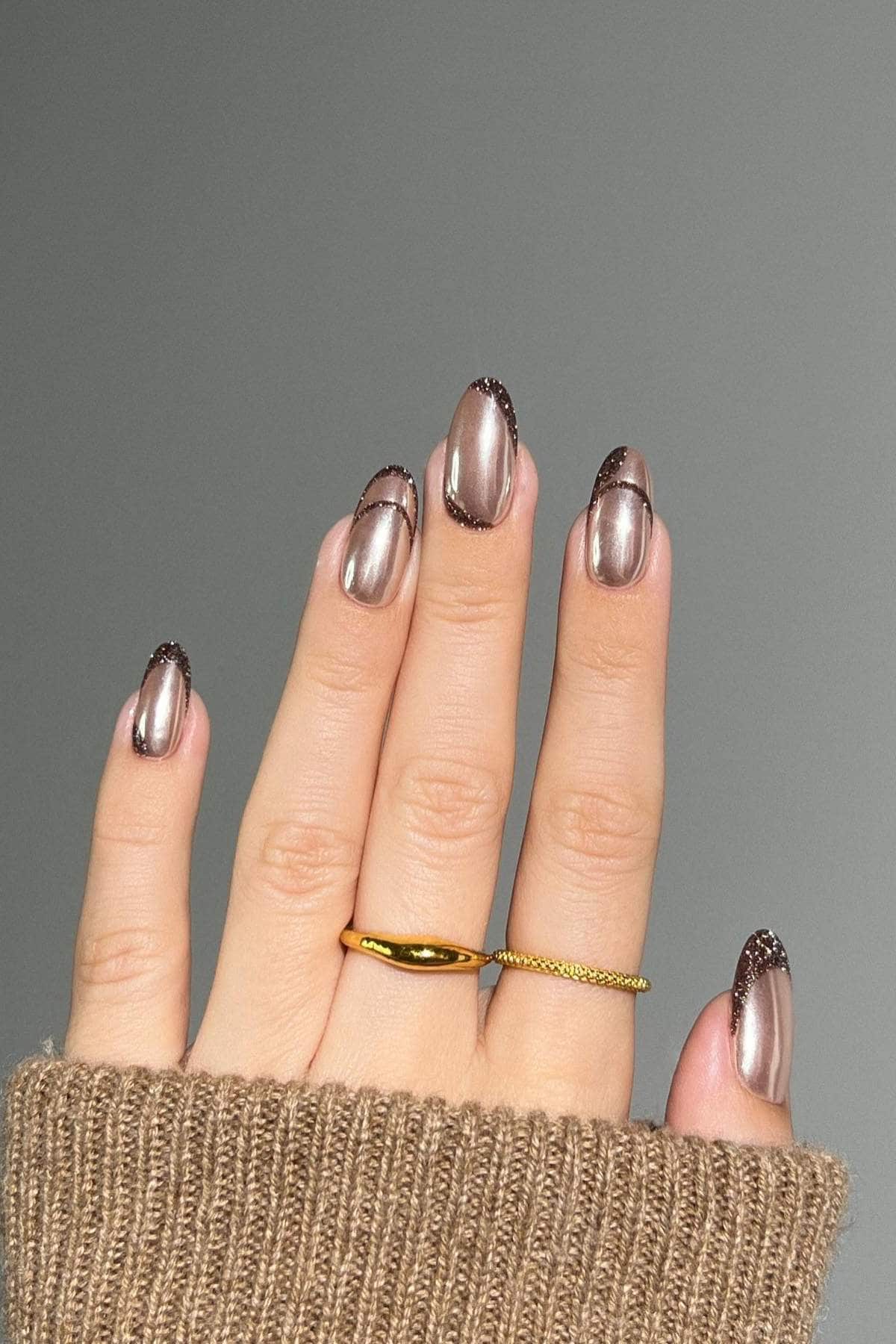 A hand with metallic nude almond-shaped nails featuring dark glitter tips and wavy lines, wearing two gold rings, against a gray background.