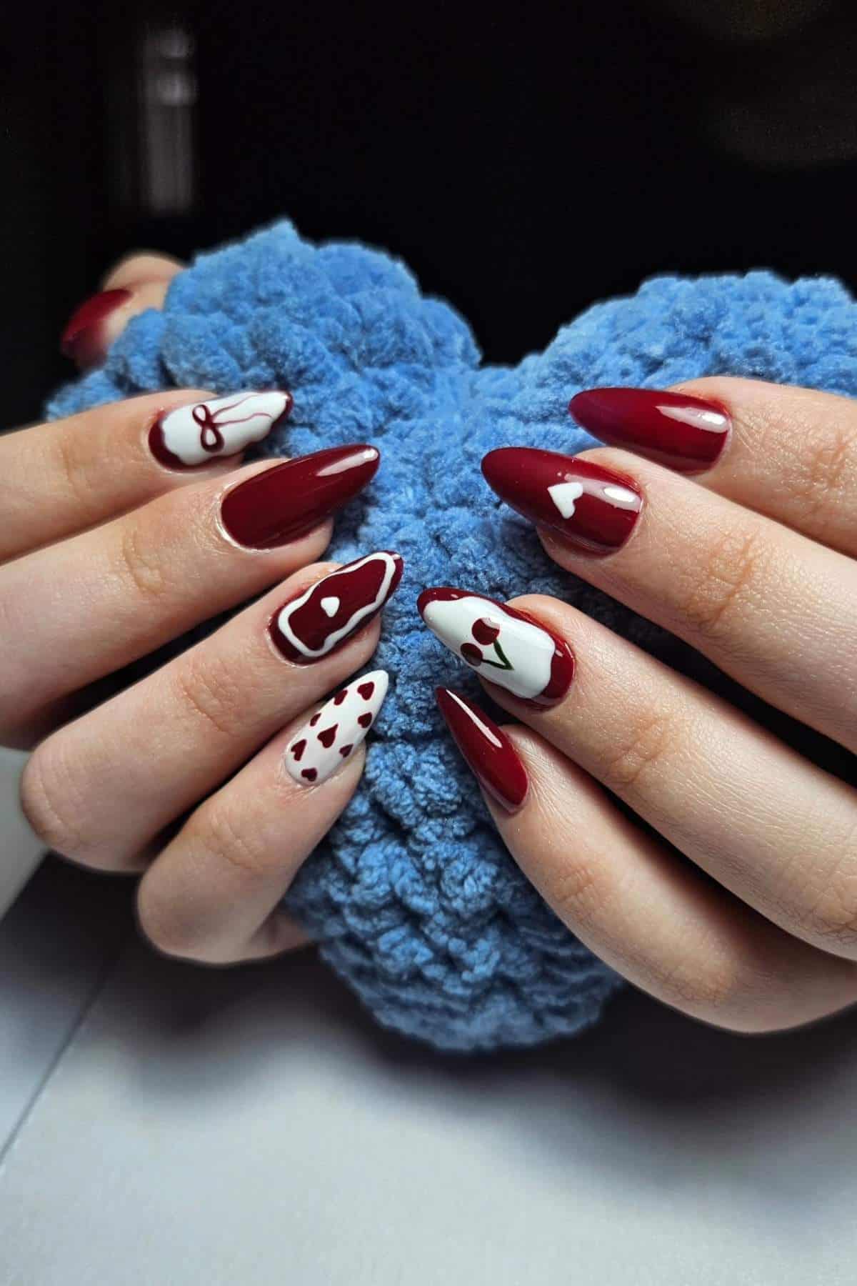 Hands with long almond-shaped nails painted in burgundy and white, featuring various heart and abstract designs, holding a blue textured object.