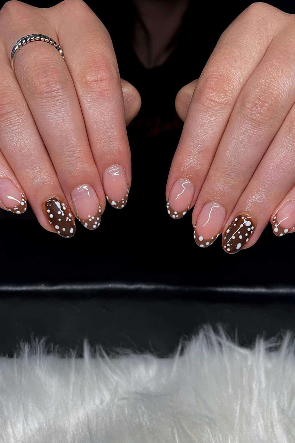 Close-up of hands with almond-shaped nails featuring brown French tips, white dots, and some clear sections; silver ring on left index finger.