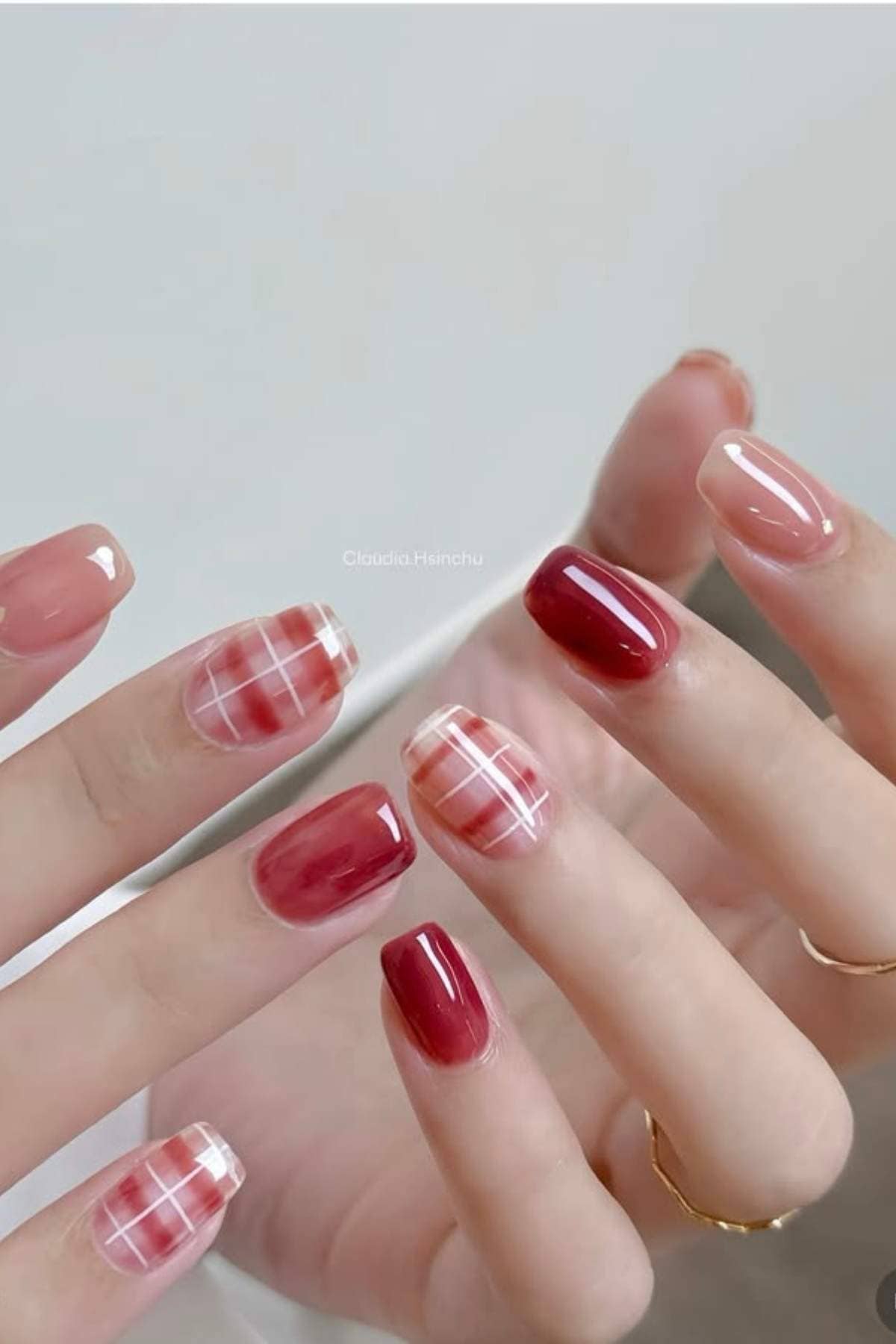 A hand with manicured nails featuring red, plaid, and nude designs. The nails are medium length and square-shaped. Two gold rings are worn on the fingers.