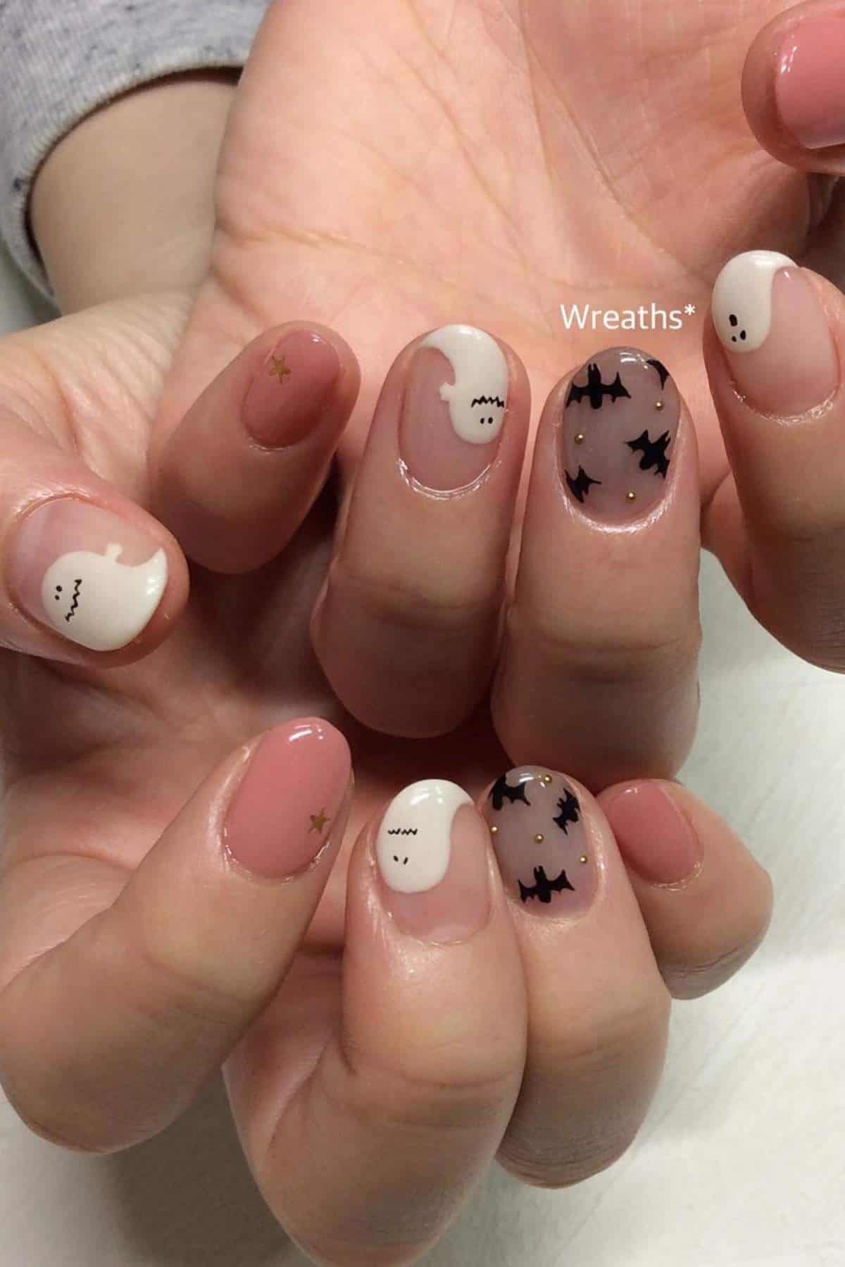 Hands with nude pink nails featuring Halloween-themed art: white ghosts and black bats, plus small gold star accents on some nails.
