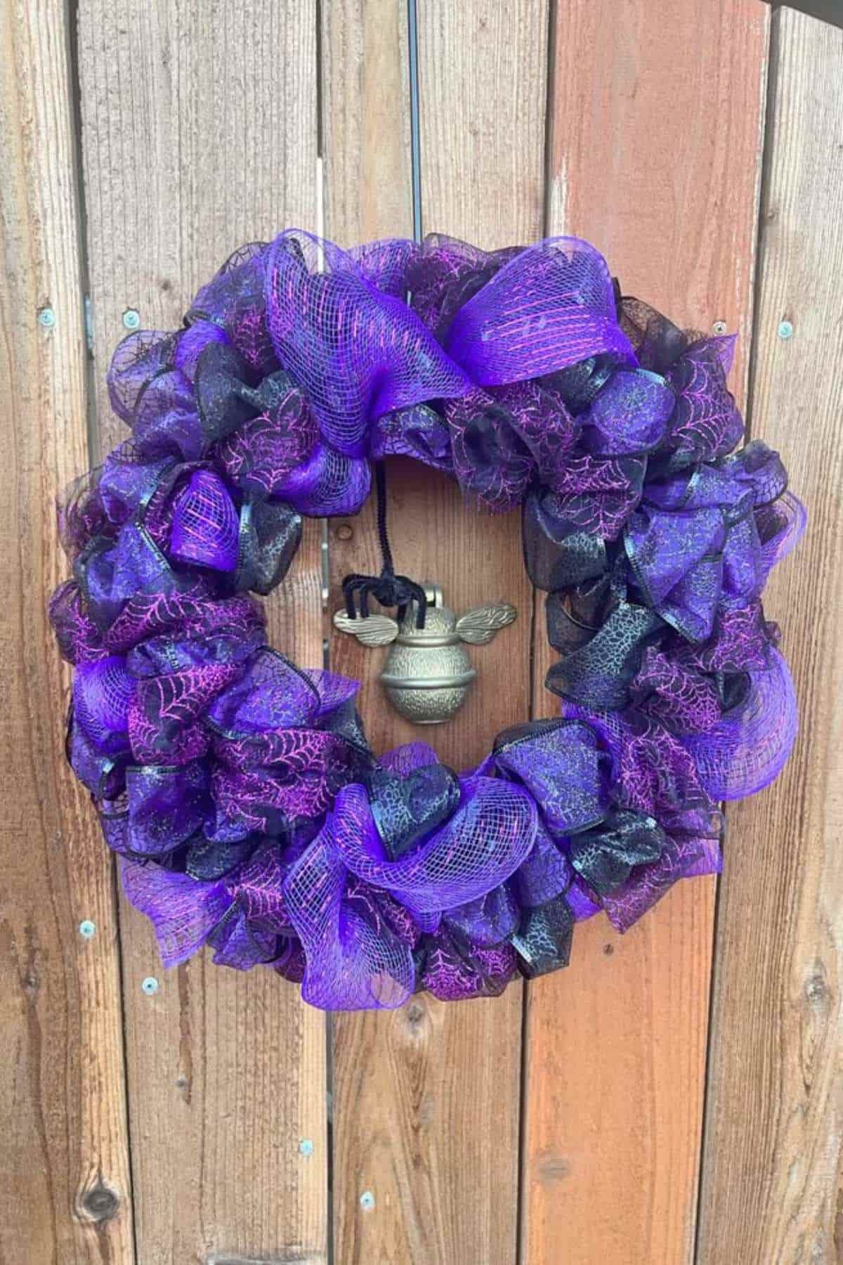 A wreath made of black and purple mesh ribbon with spiderweb patterns, featuring a small cauldron decoration hanging in the center, displayed on a wooden door.
