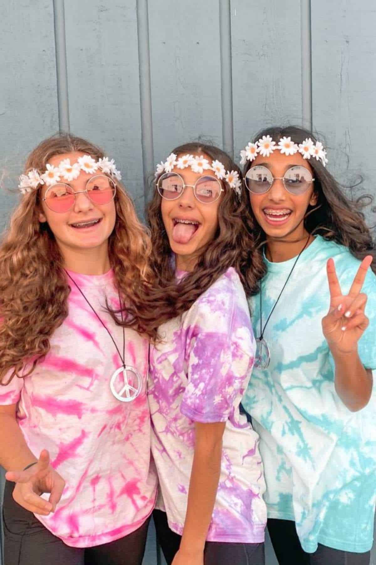Three girls wearing tie-dye shirts, flower headbands, peace sign necklaces, and round glasses stand in front of a gray wall, smiling and making peace signs.