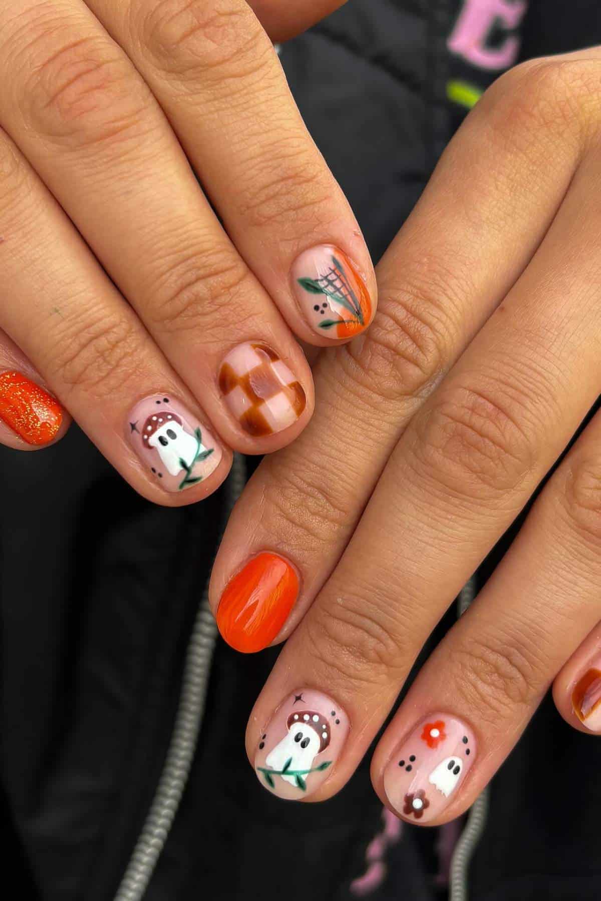 Hands with orange, plaid, and ghost-themed nail art, including flowers and abstract designs, against a dark background.