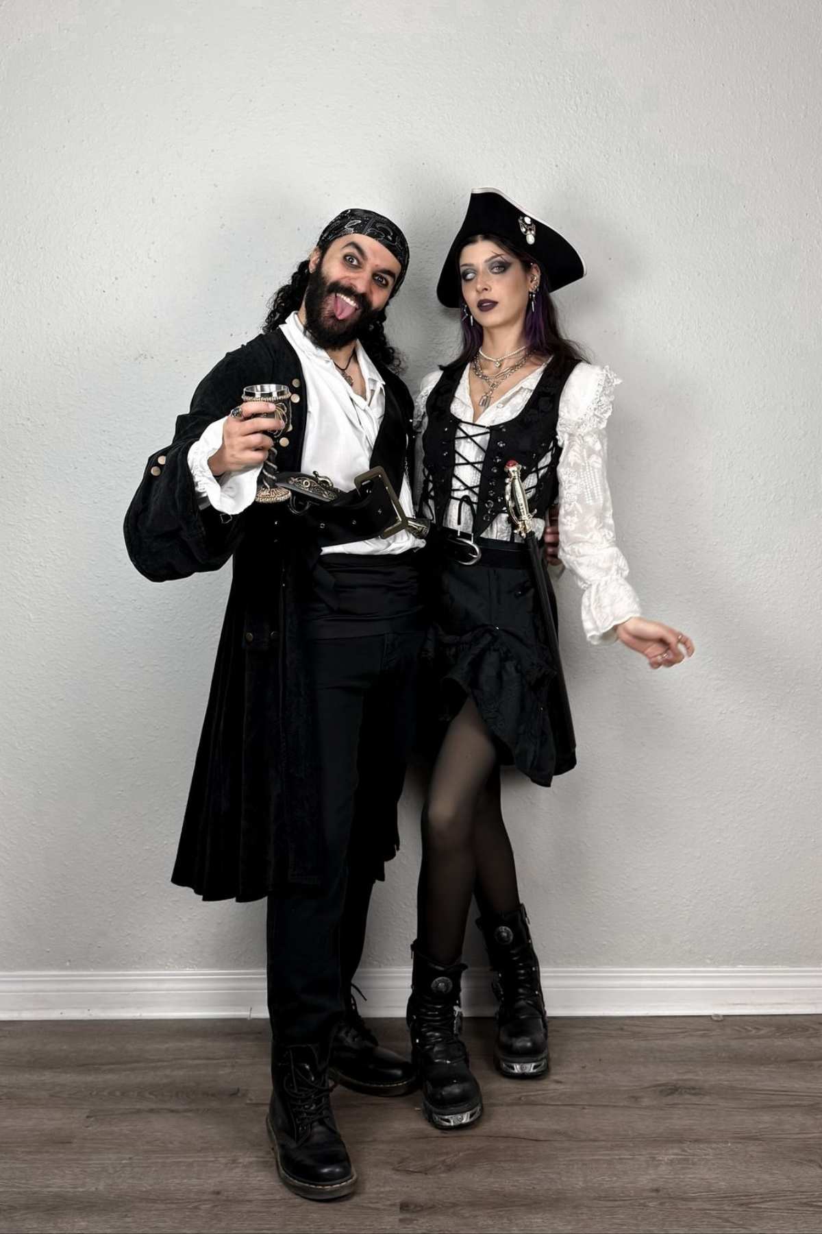 Two people dressed in black and white pirate costumes stand together indoors, one holding a drink and making a playful face, both wearing boots and standing on a wooden floor against a plain wall.