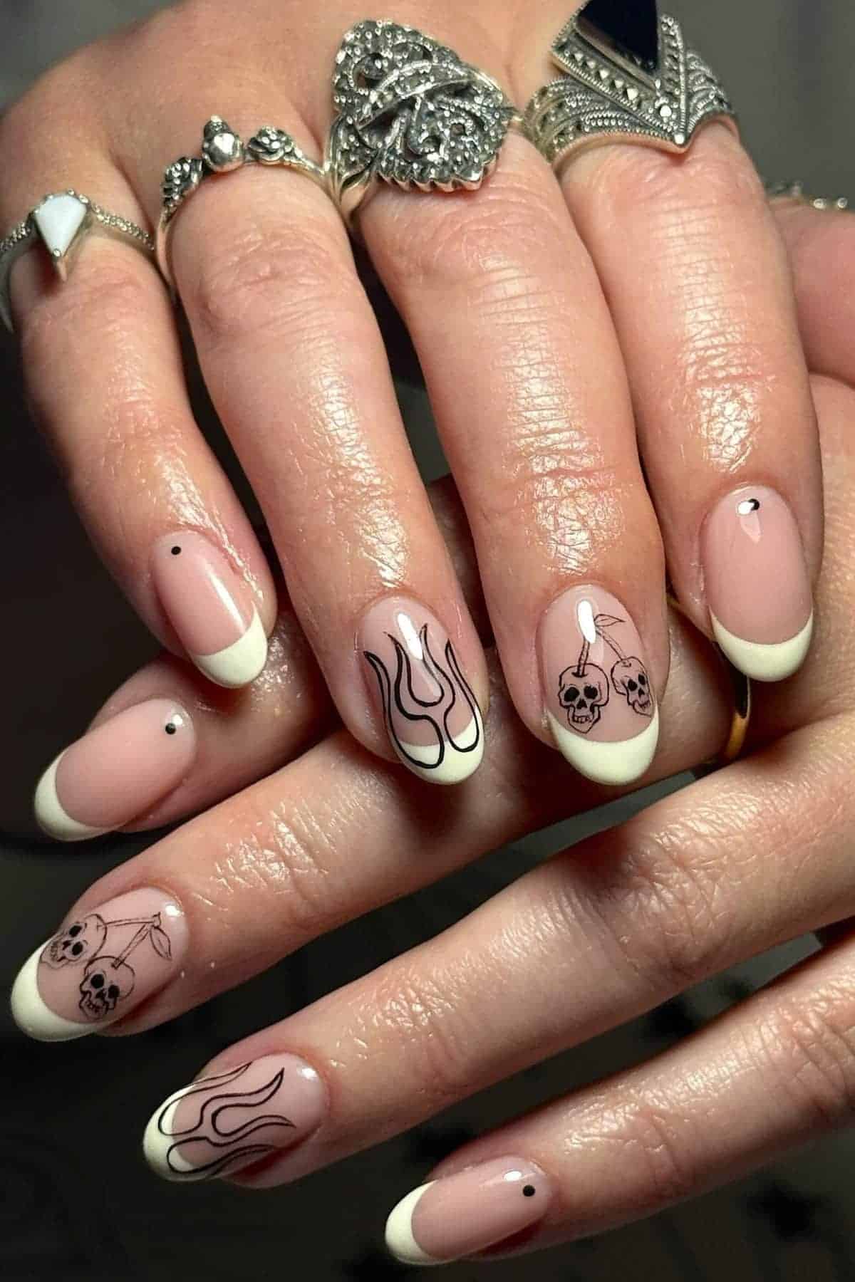 Hands with almond-shaped nails featuring white French tips, black flame and skull nail art, and small black dot accents. Fingers are adorned with multiple silver rings.