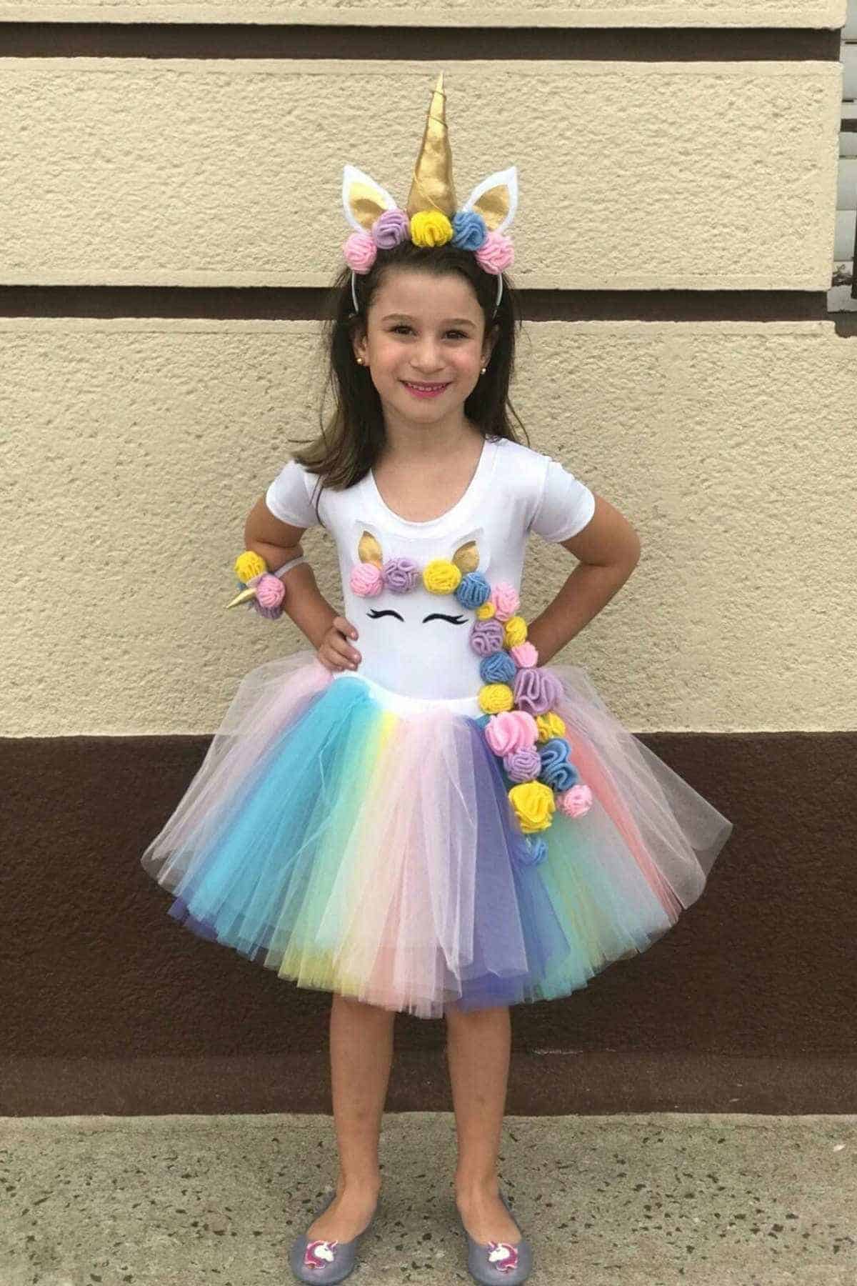Young girl stands smiling in a unicorn-themed costume with a rainbow tutu, floral decorations, and a unicorn horn headband in front of a textured wall.