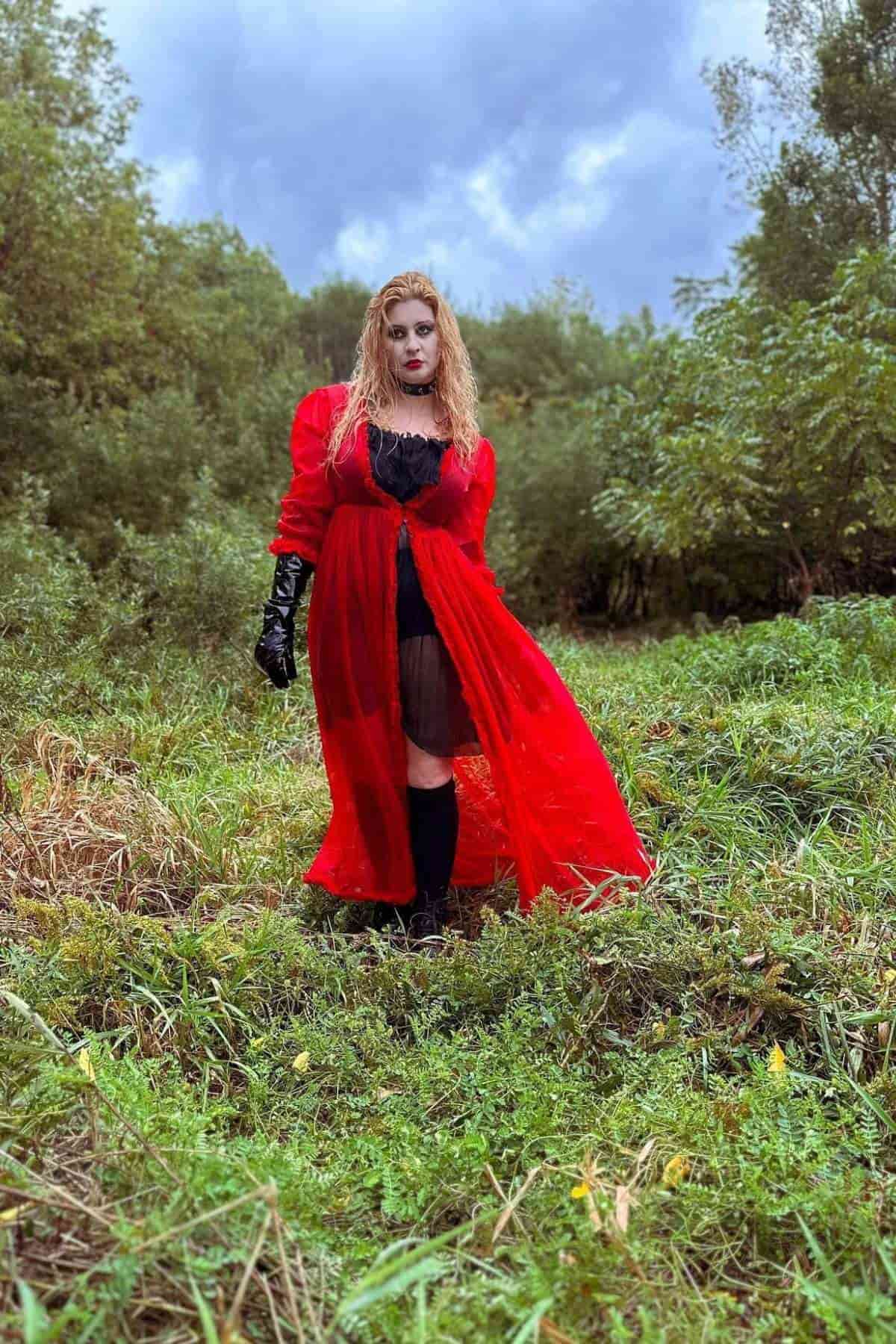 A person wearing a sheer red dress, black gloves, and boots stands on grass in front of dense green foliage under a cloudy sky.
