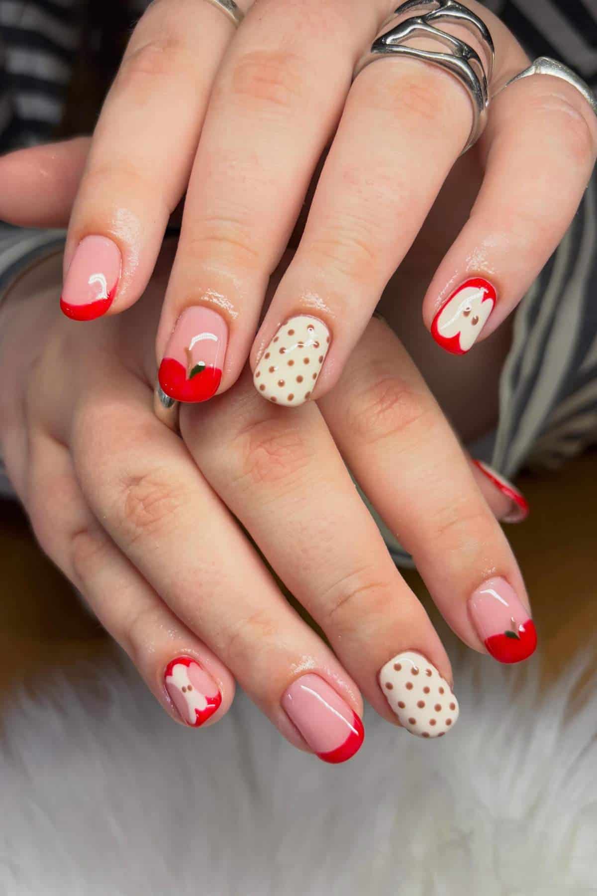 Hands with manicured nails featuring red tips, cherry designs, white nails with red polka dots, and some with small cherry details. The person is wearing silver rings.