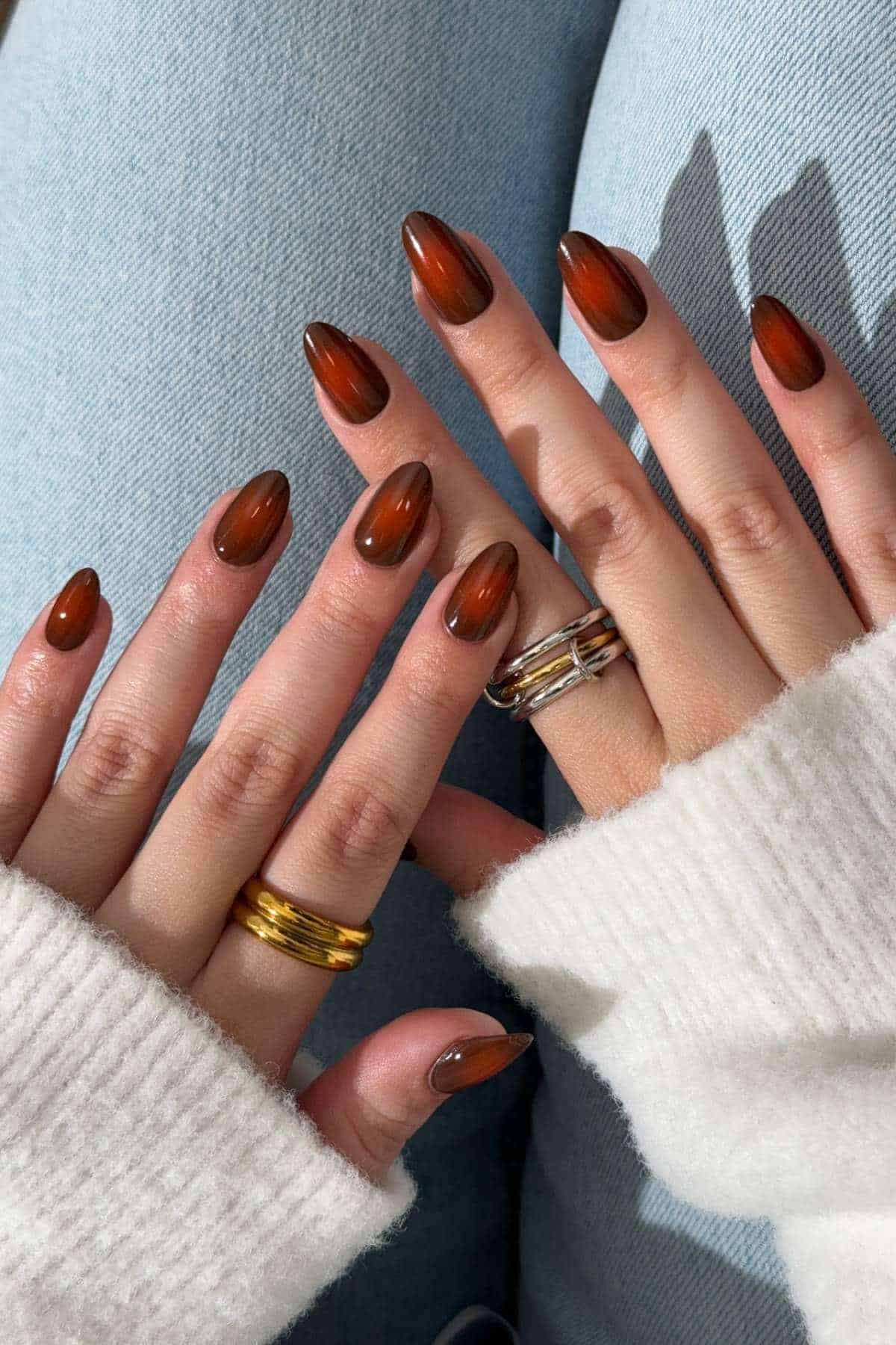 Close-up of hands with almond-shaped, brown gradient nail polish, wearing gold and silver rings, and resting on light blue jeans.