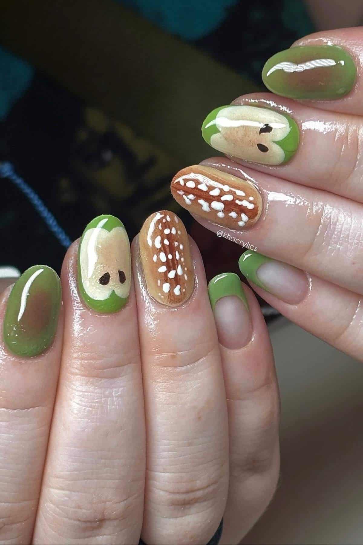 Close-up of hands with green and brown nail art resembling apple and one seed with white spots, on rounded fingernails.