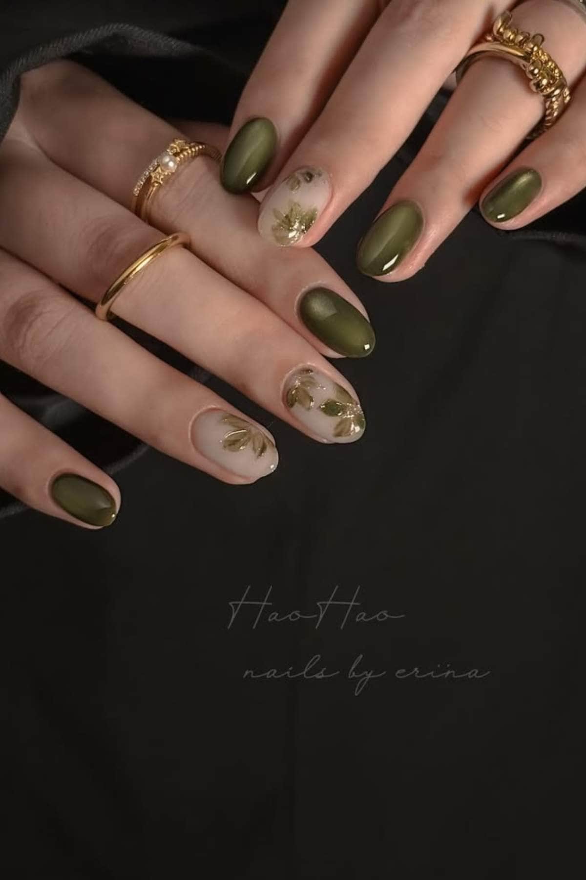 Close-up of hands with dark green and clear nails featuring gold leaf accents, accessorized with gold rings.