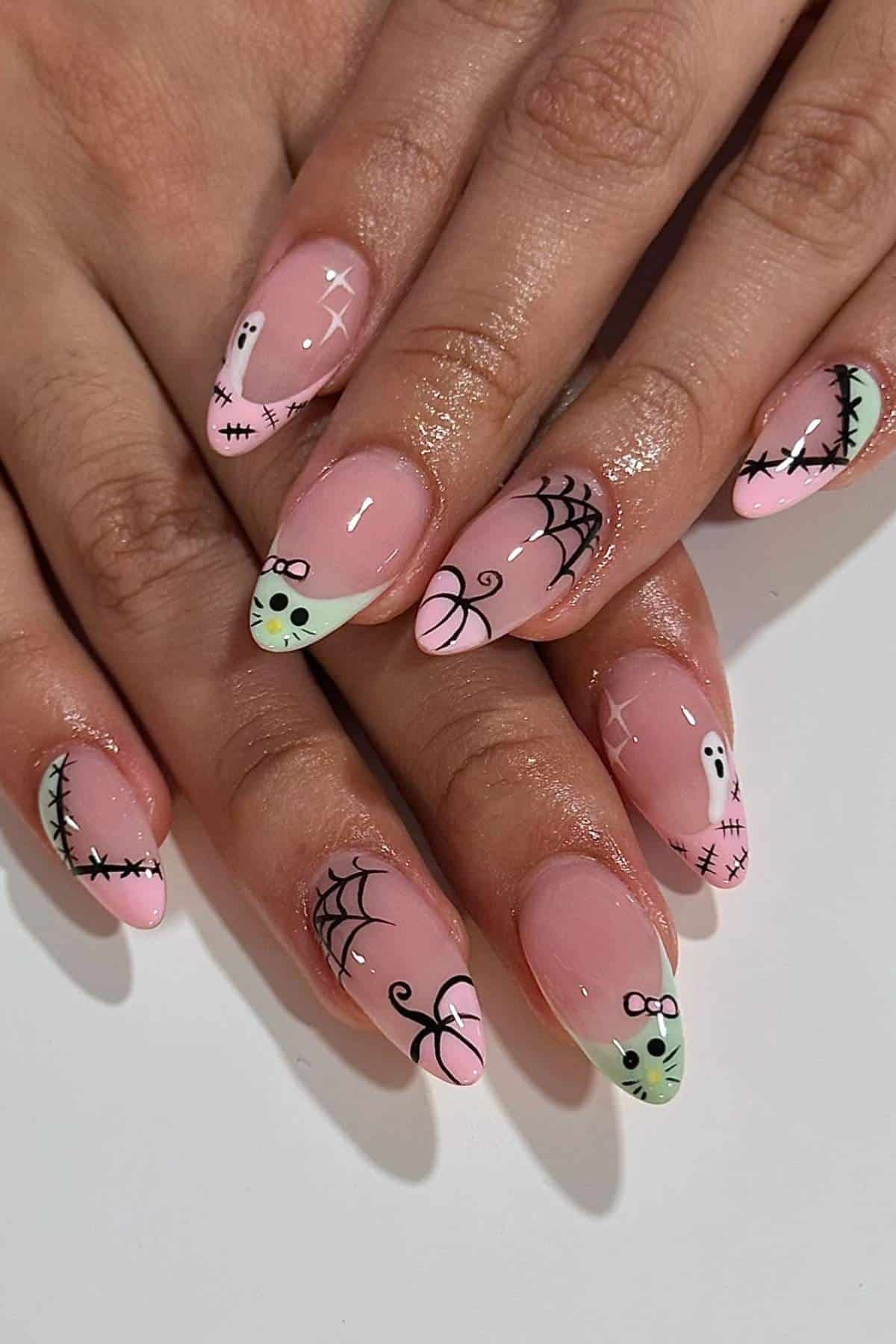 Hands with almond-shaped nails featuring pink and green tips, decorated with black spiderwebs, stitches, ghosts, and small face designs on a glossy finish.