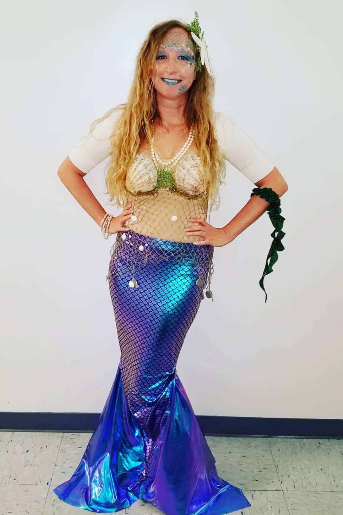 Person dressed in a shiny blue mermaid costume with a fishtail skirt, seashell top, netting, and aquatic accessories, standing against a plain wall.