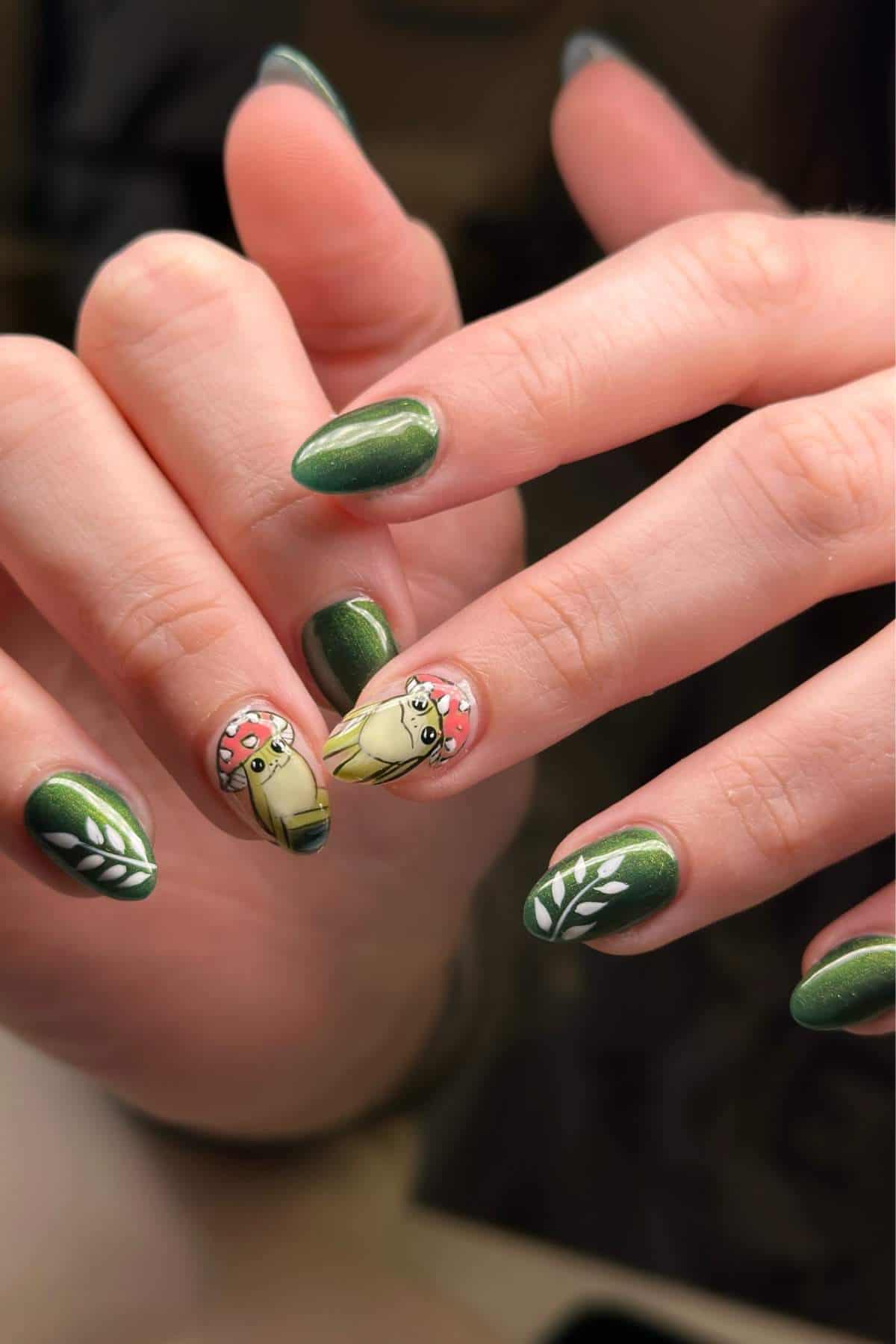 Hands with green manicure; ring and middle fingernails display cartoon mushrooms with faces, other nails have white leaf designs on a green background.