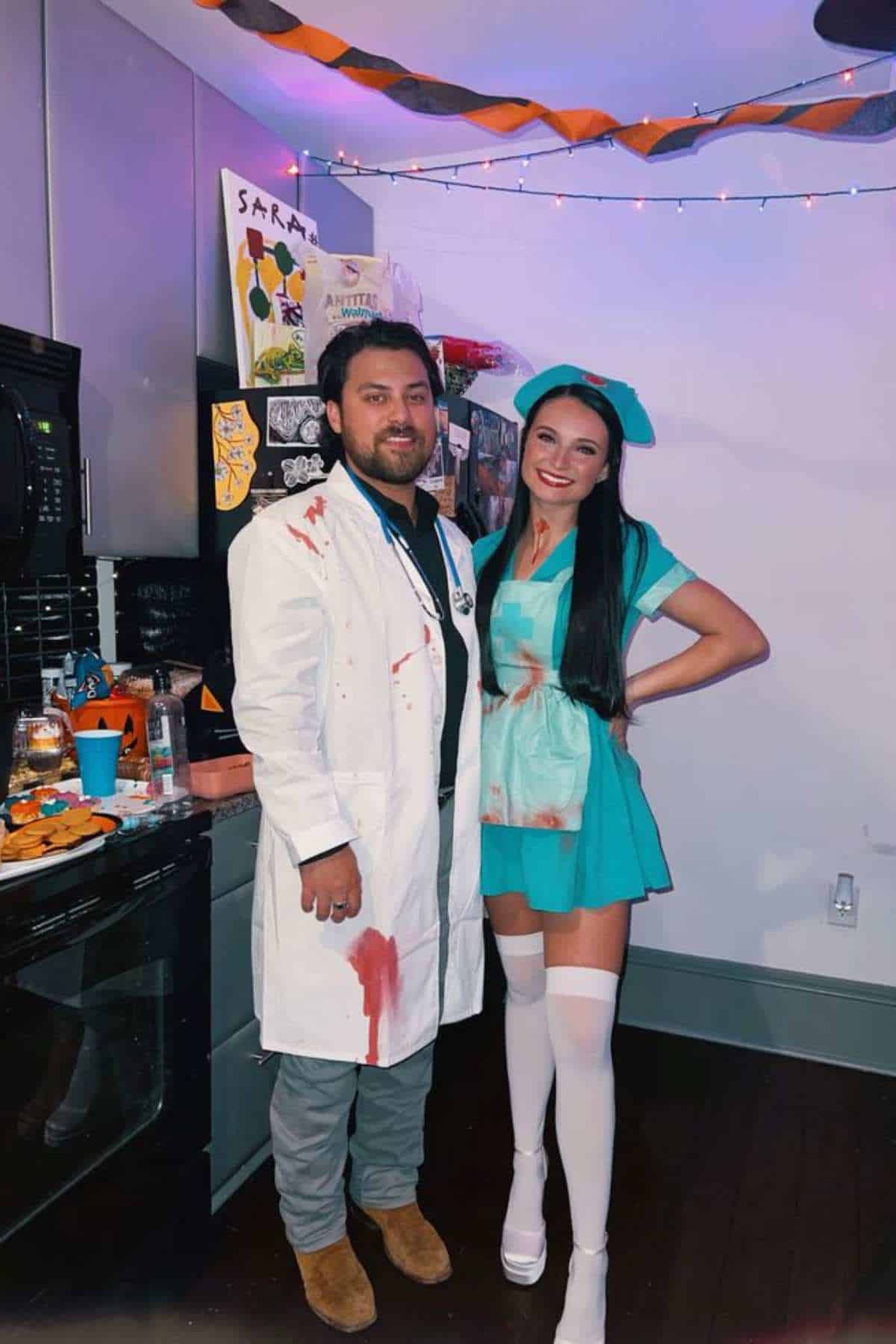 Two people in a kitchen wear matching blood-splattered costumes; one is dressed as a doctor, the other as a nurse. Halloween decorations and party food are visible around them.