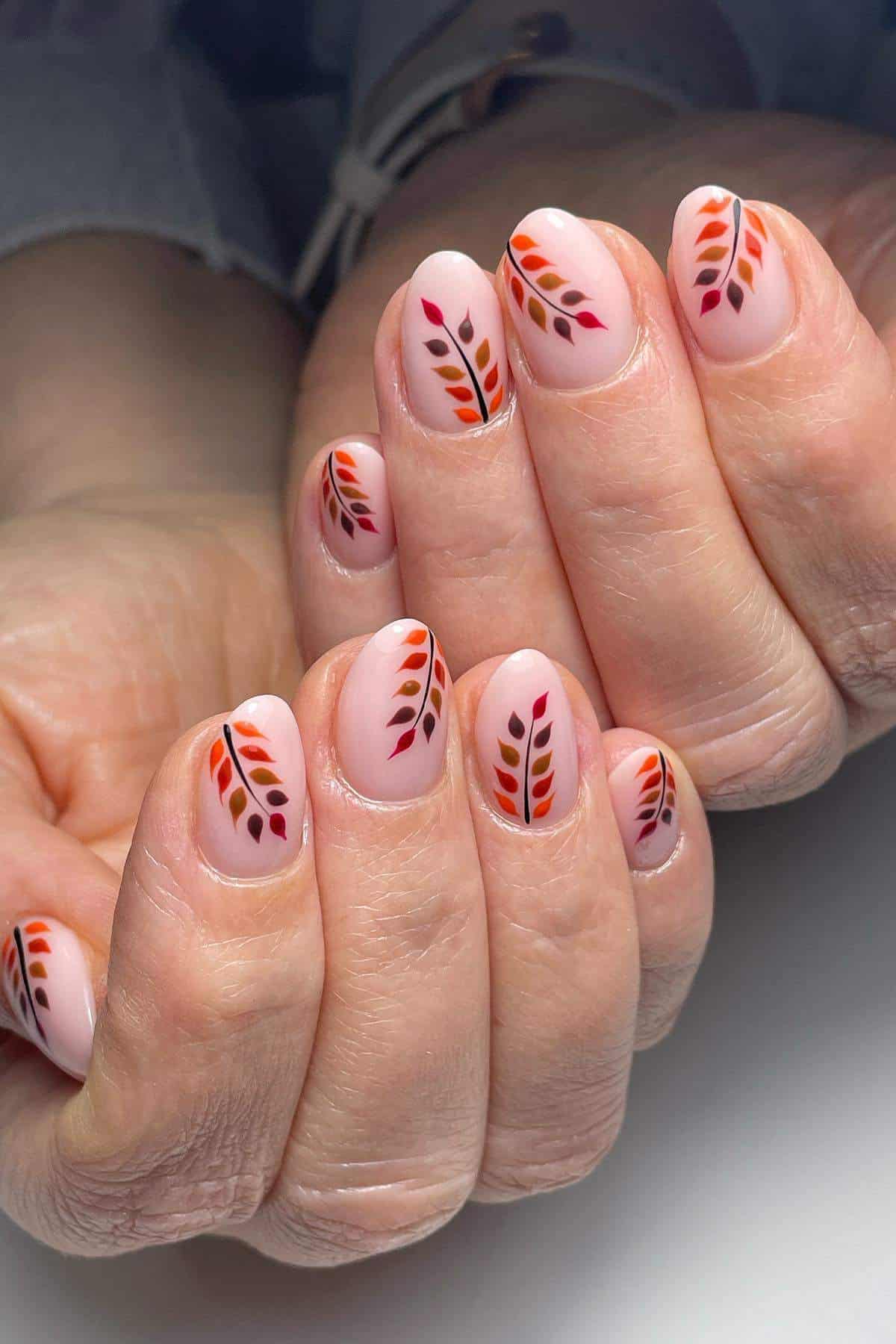 Close-up of hands with light pink nail polish featuring leaf-like designs in orange, red, and brown on each fingernail.