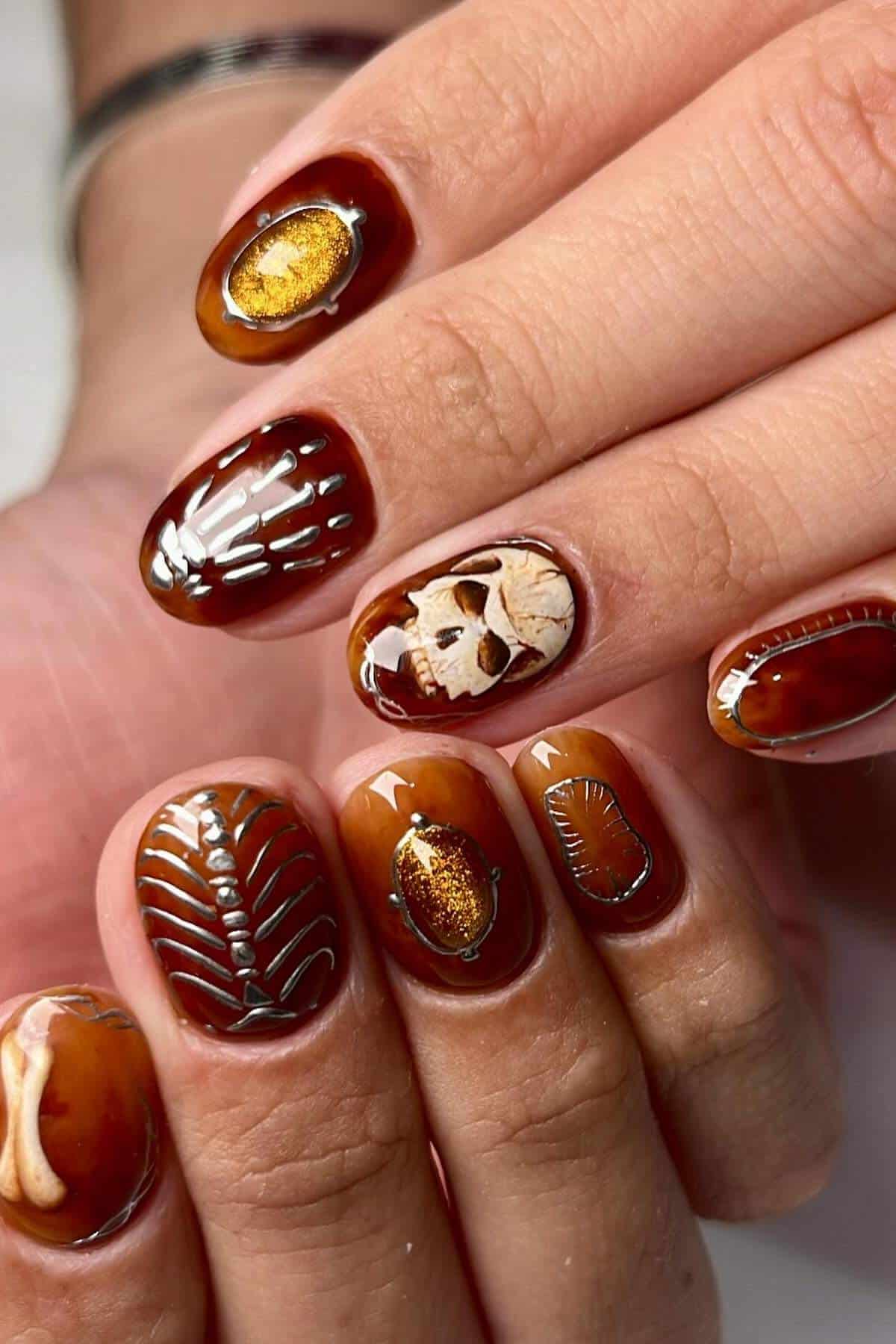Close-up of a hand with brown fossil-inspired nail art, featuring gold accents, a skull, bones, and leaf designs on each fingernail.