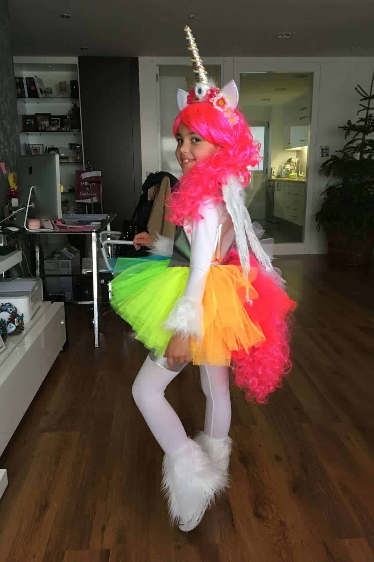 Child in a unicorn costume with a pink wig, white horn, colorful tutu, wings, and furry boots, standing in a modern living room.