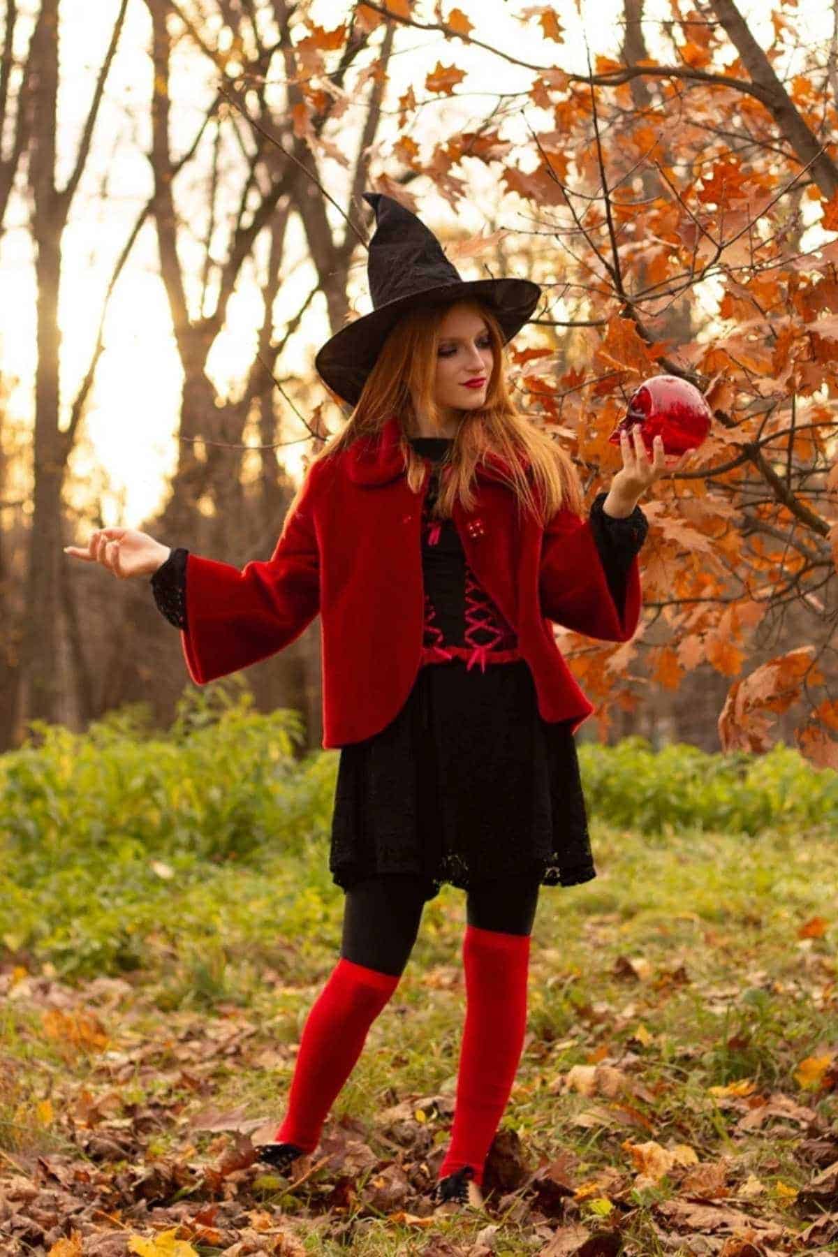 Woman in a witch costume with a red jacket and hat stands outdoors on fallen leaves, holding a red apple, surrounded by autumn trees.