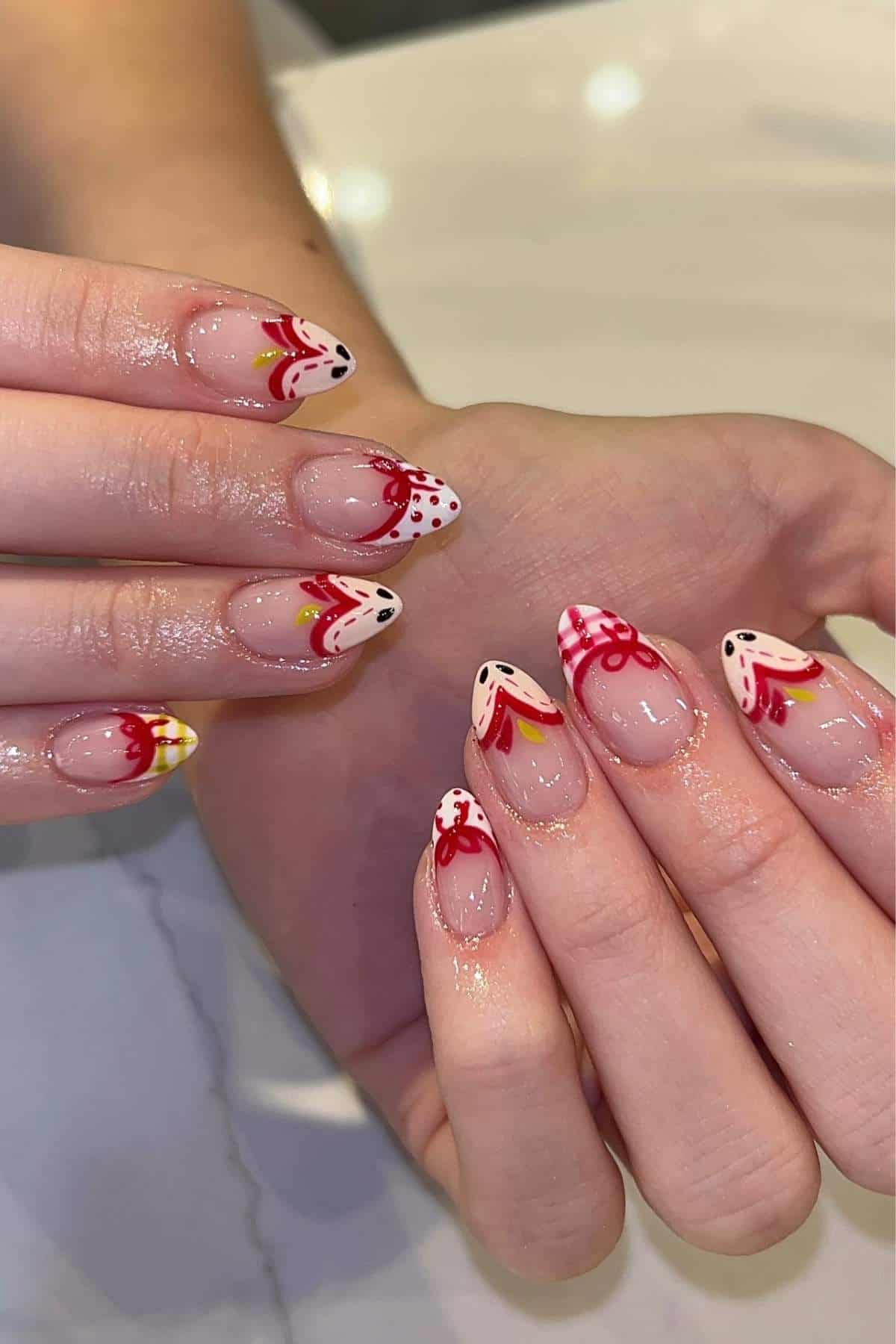 Hands with almond-shaped nails featuring red, yellow, and white floral and abstract designs on a clear base. Nails and fingers appear glossy, with a marble surface in the background.