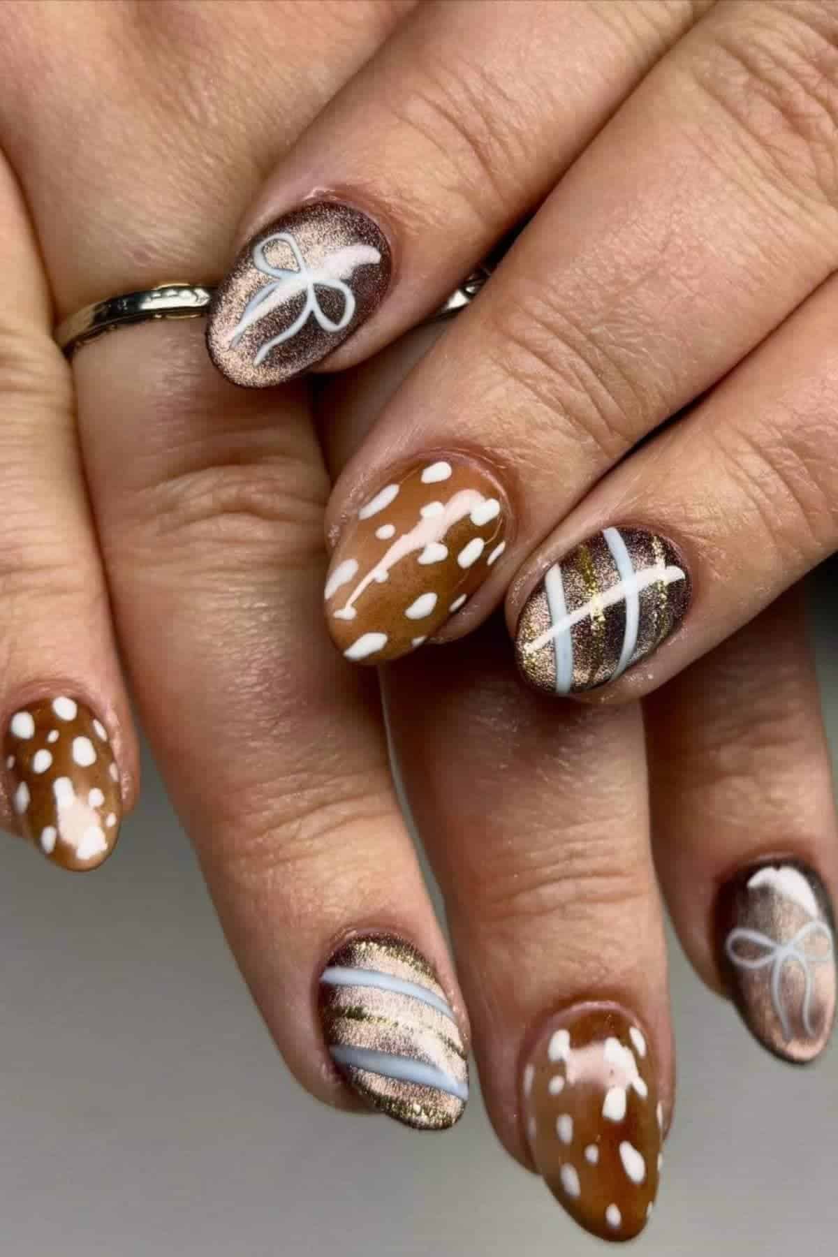 Close-up of hands with brown and gold nail art featuring white polka dots, lines, and bow designs. Nails are almond-shaped and polished with glossy finish.