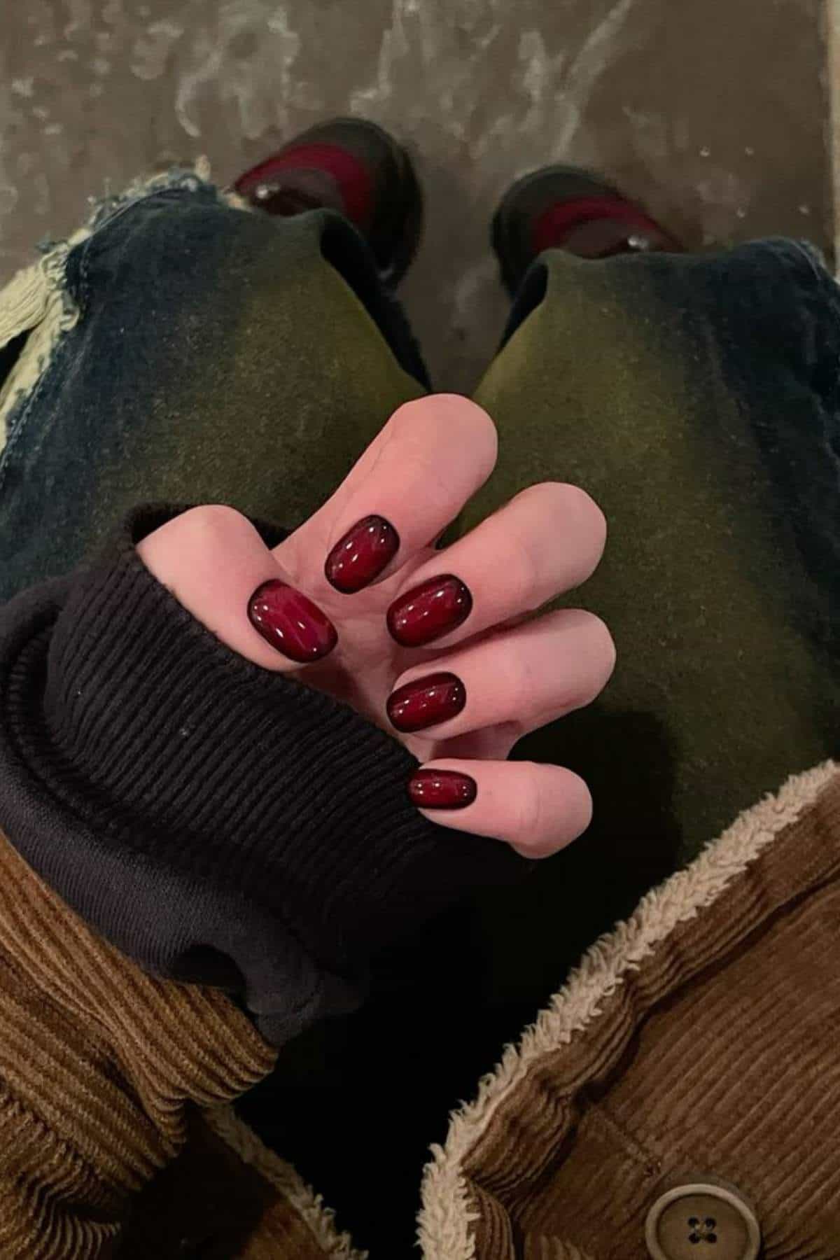 A person with dark red oval-shaped manicured nails wears a brown corduroy jacket and dark jeans with ripped knees, sitting down.