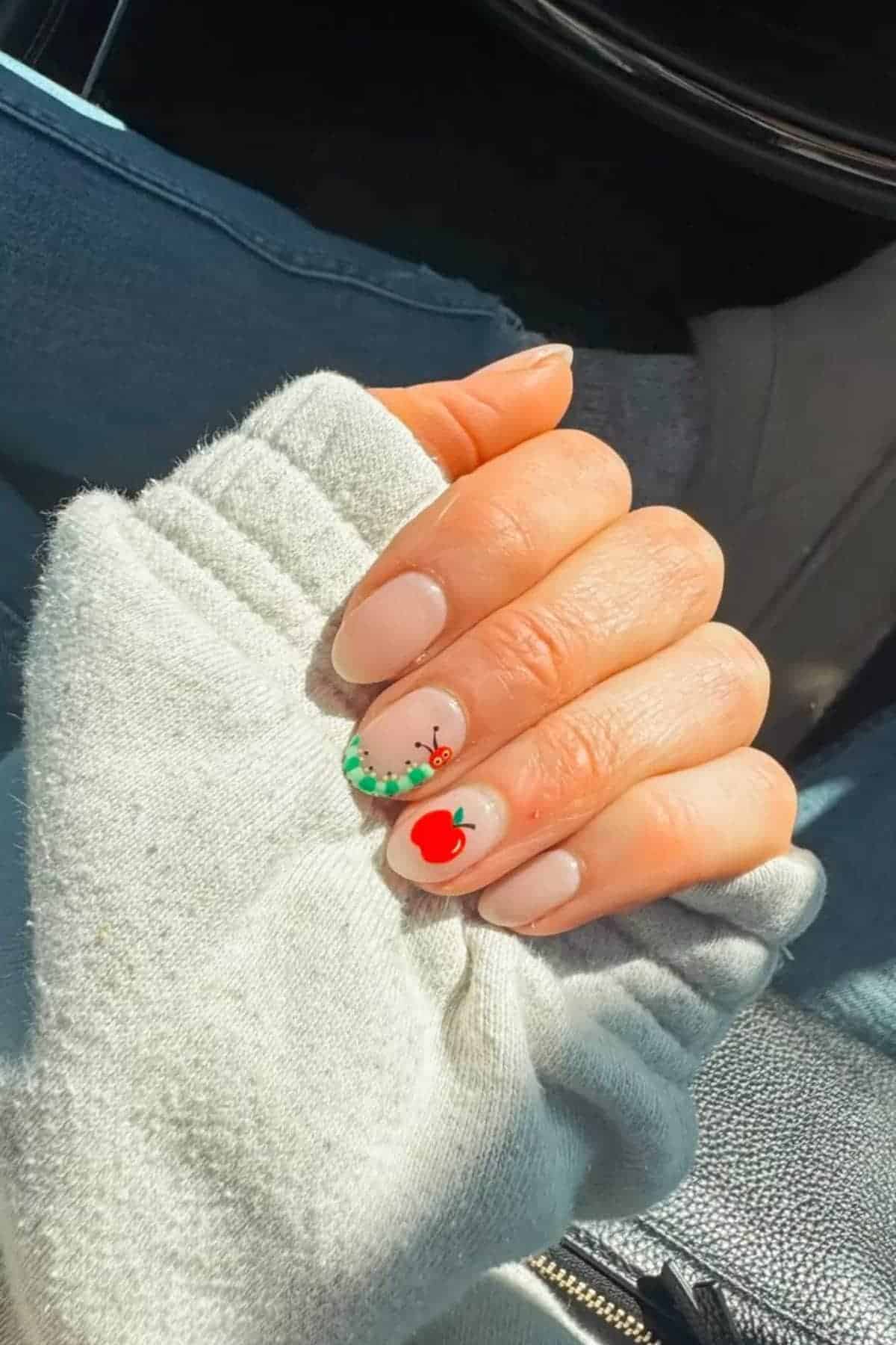 A hand with light nude nail polish; two nails feature cherry nail art and a green bead-like accent. The person is wearing a light-colored sweater and blue jeans.