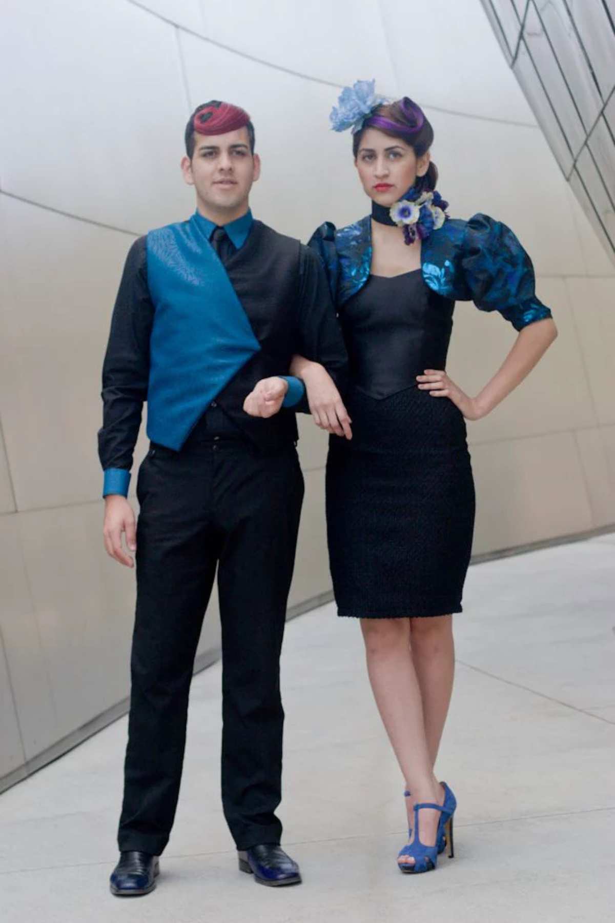A man and a woman pose side by side in coordinated blue and black formal outfits with matching hats; the woman also wears blue flowers in her hair.