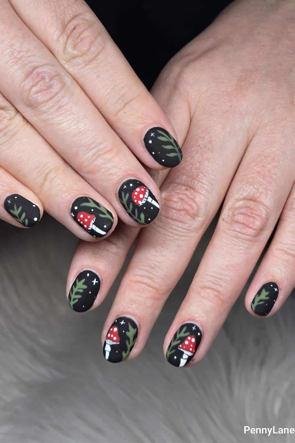 Hands with matte black nail polish featuring red mushrooms, green leaves, and small white stars as nail art designs.