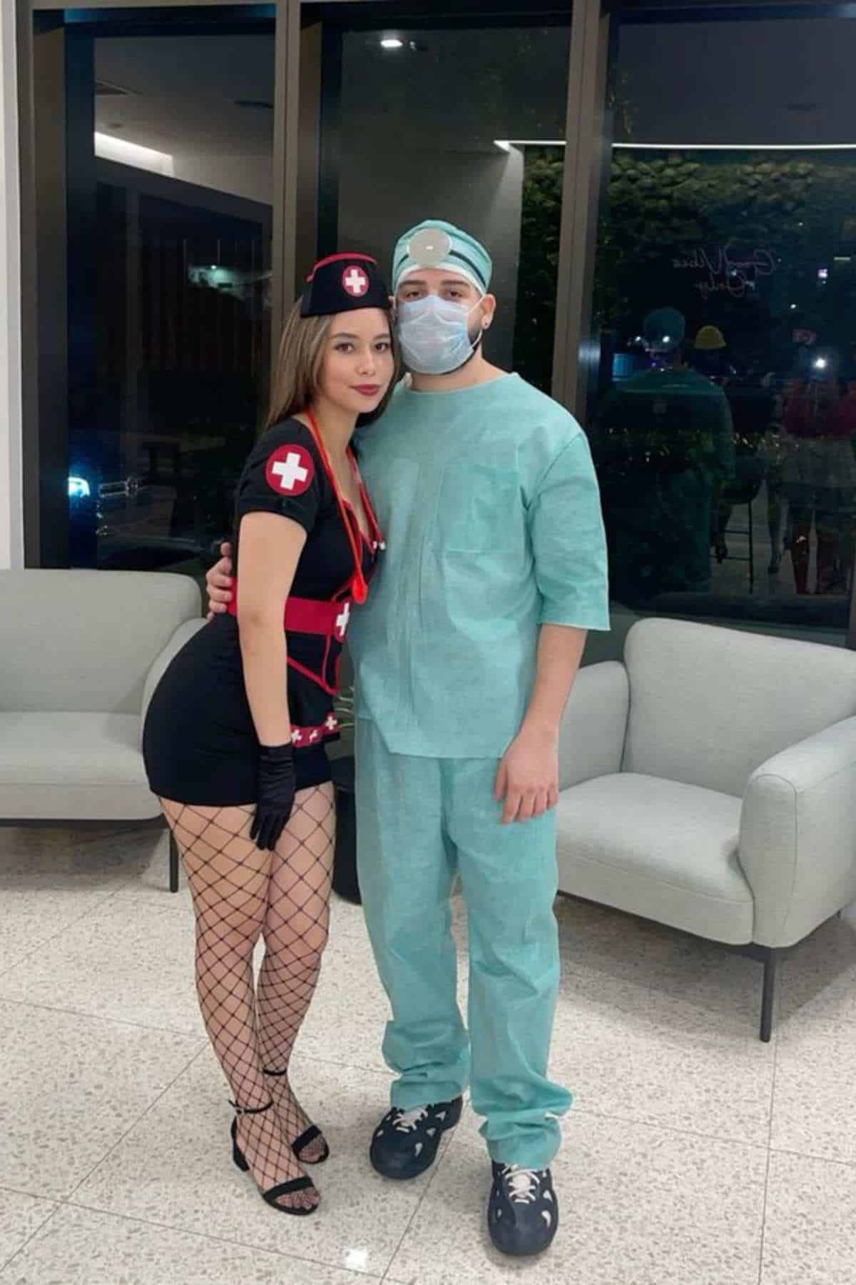 Two people pose indoors; one wears a nurse costume with fishnet stockings, and the other wears green medical scrubs, a mask, and a cap.