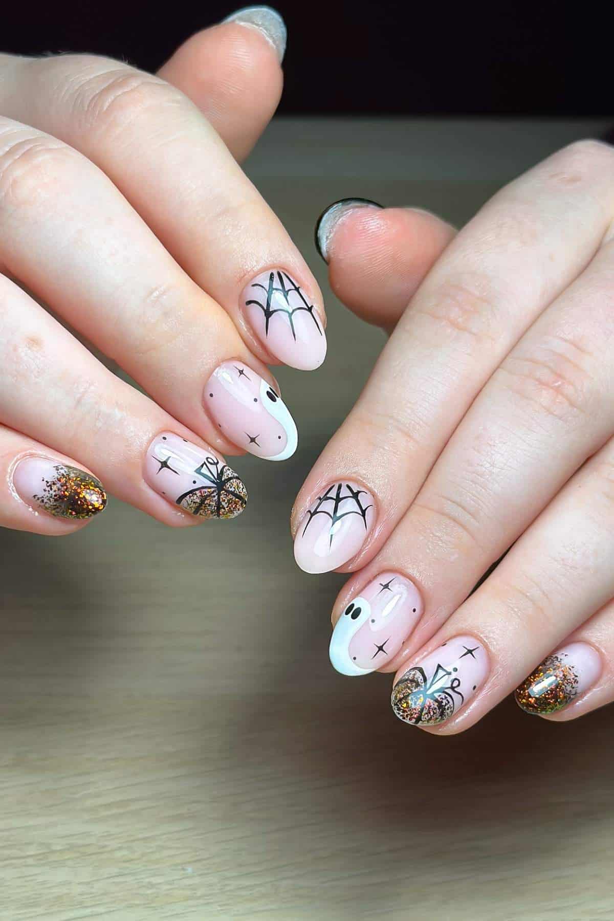 Hands with Halloween-themed nail art featuring spiderwebs, a ghost, stars, and glittery gold tips on a natural pink base, displayed on a wooden surface.