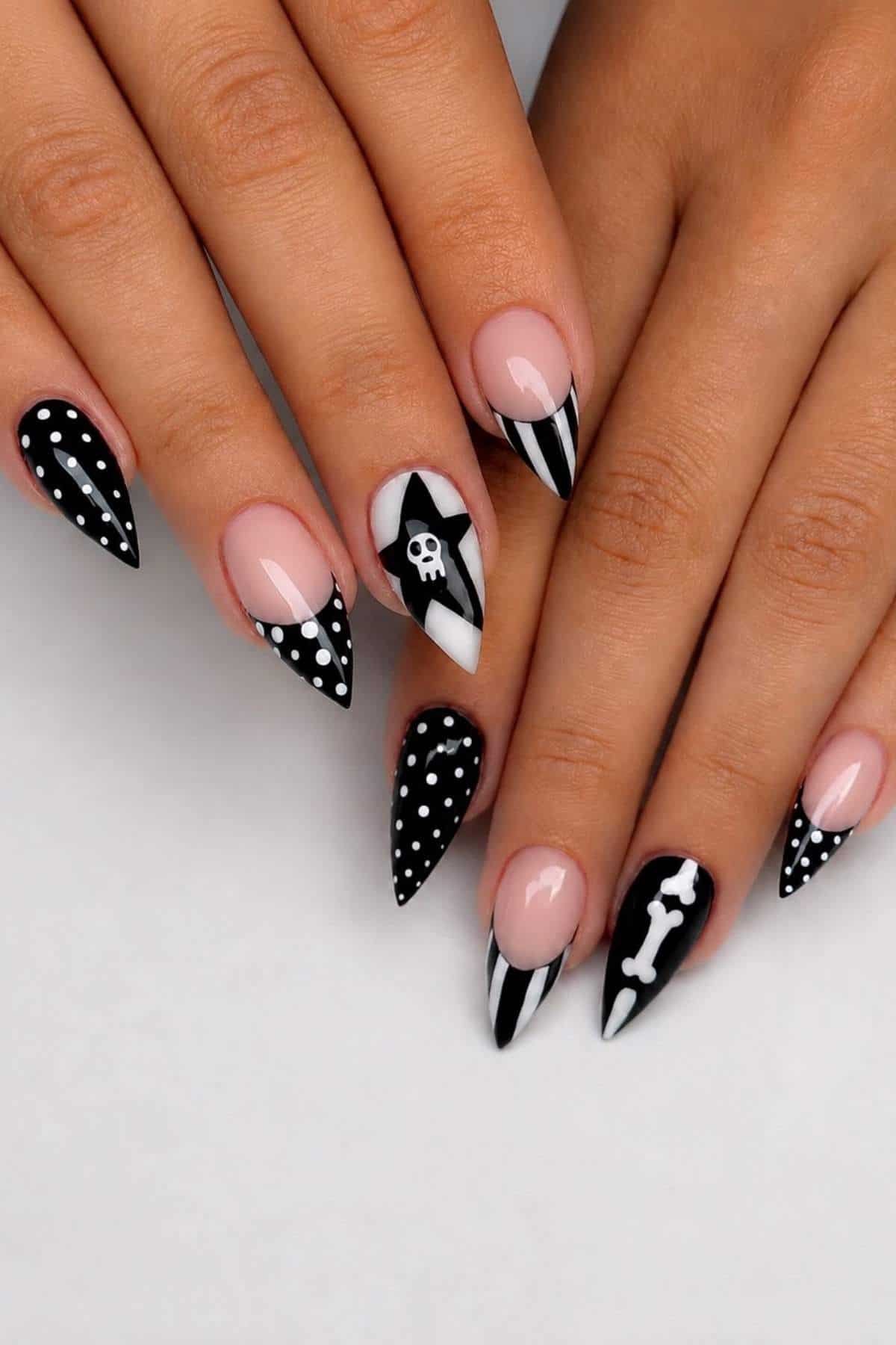 Hands with long, pointed nails featuring black and white Halloween-themed designs, including stripes, polka dots, a skull, and a bone on a white background.