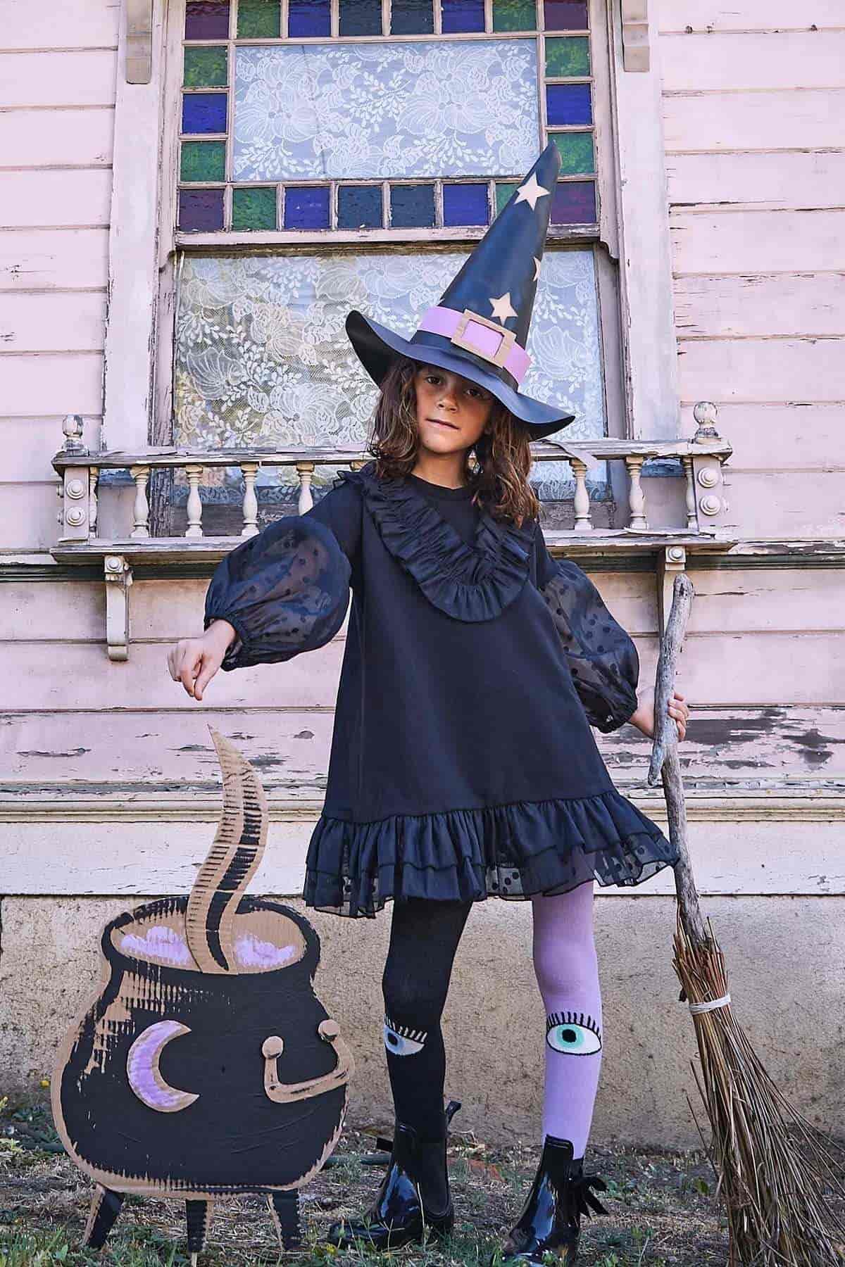 Child in a black witch costume and hat stands by a broom and a cardboard cauldron prop in front of an old house with a stained glass window.