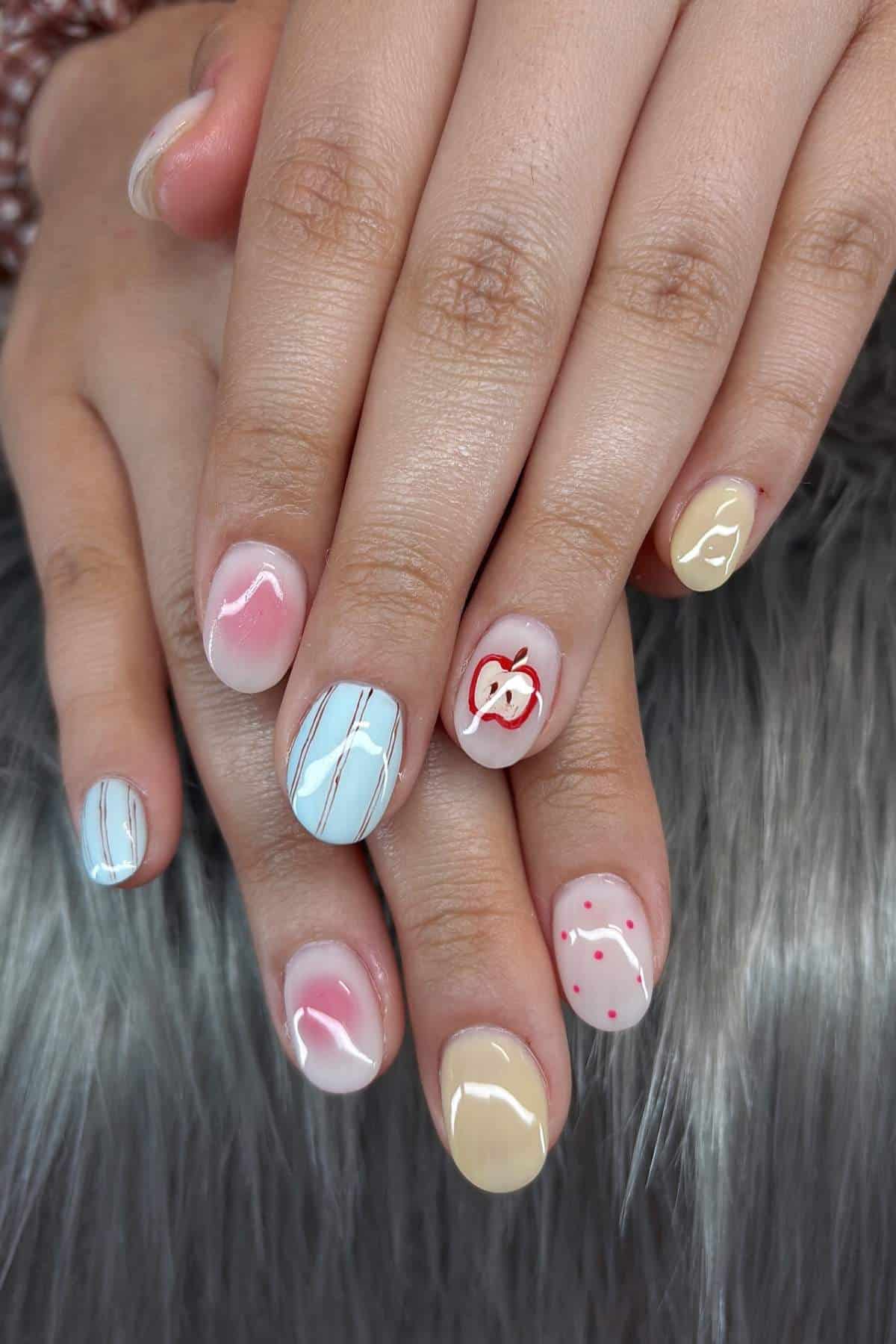 A hand with short, rounded nails features various nail art designs, including stripes, an apple, polka dots, and solid pastel colors.